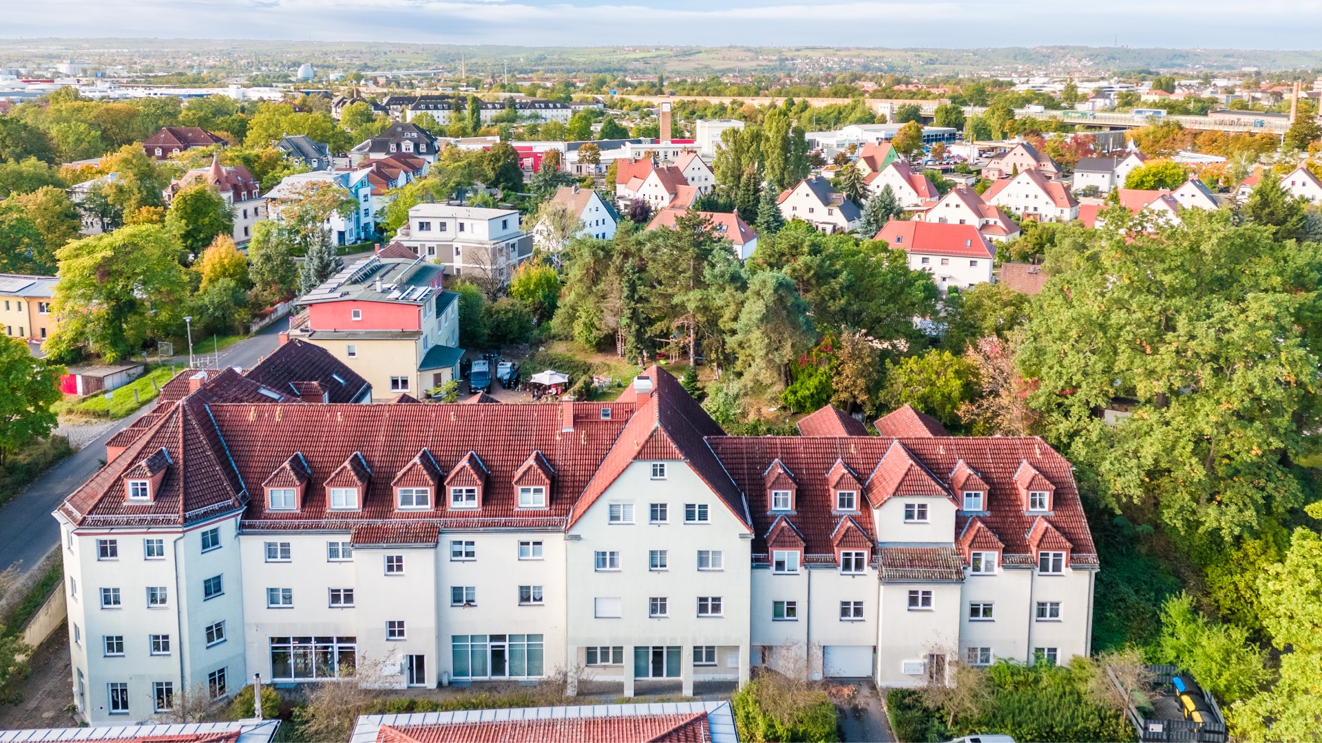 Großes Wohnhaus mit roten Dächern, umgeben von Bäumen und weiteren Häusern im Hintergrund.