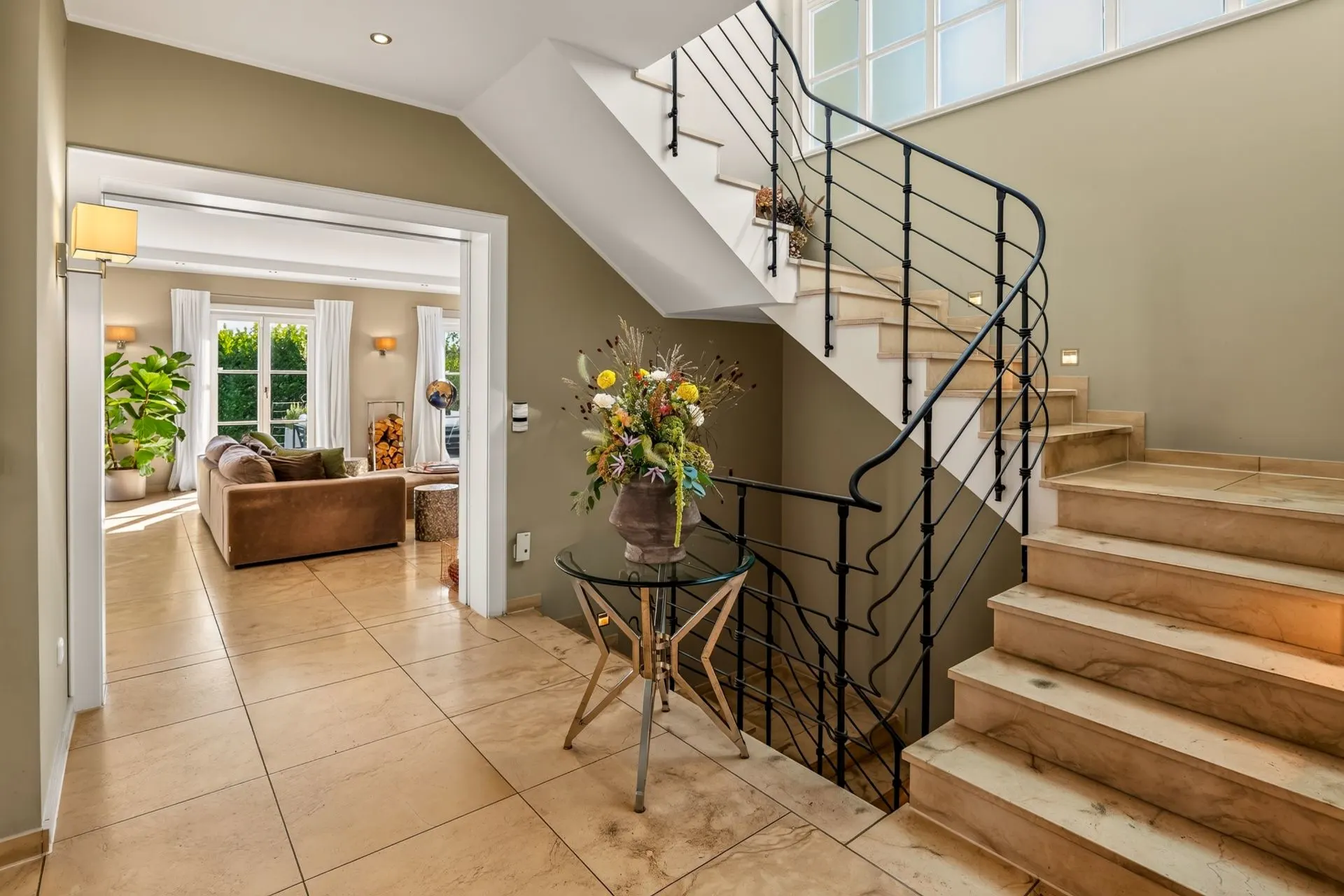 Heller Eingangsbereich mit Treppe, Glas-Tisch und Blumen. Wohnbereich mit Sofa im Hintergrund.