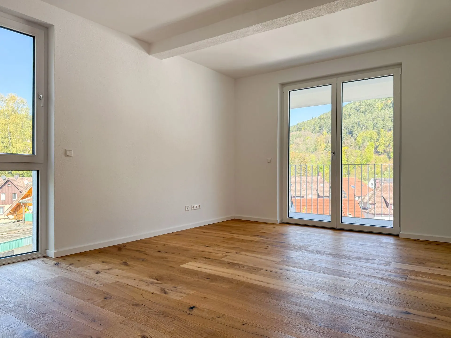 Leeres Zimmer mit Holzfußboden, großen Fenstern und Blick ins Grüne.