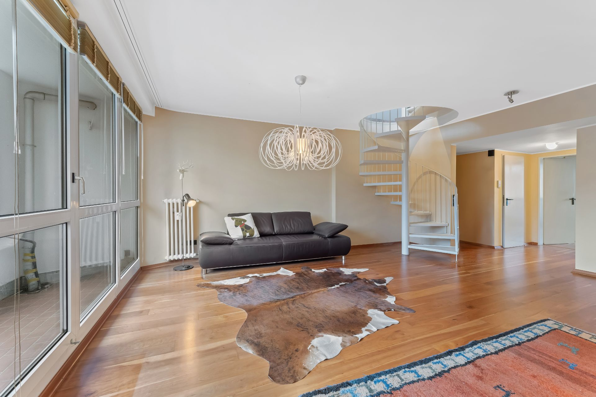 Helles Wohnzimmer mit großer Fensterfront, Ledersofa, Kuhfell-Teppich und weißer Wendeltreppe.