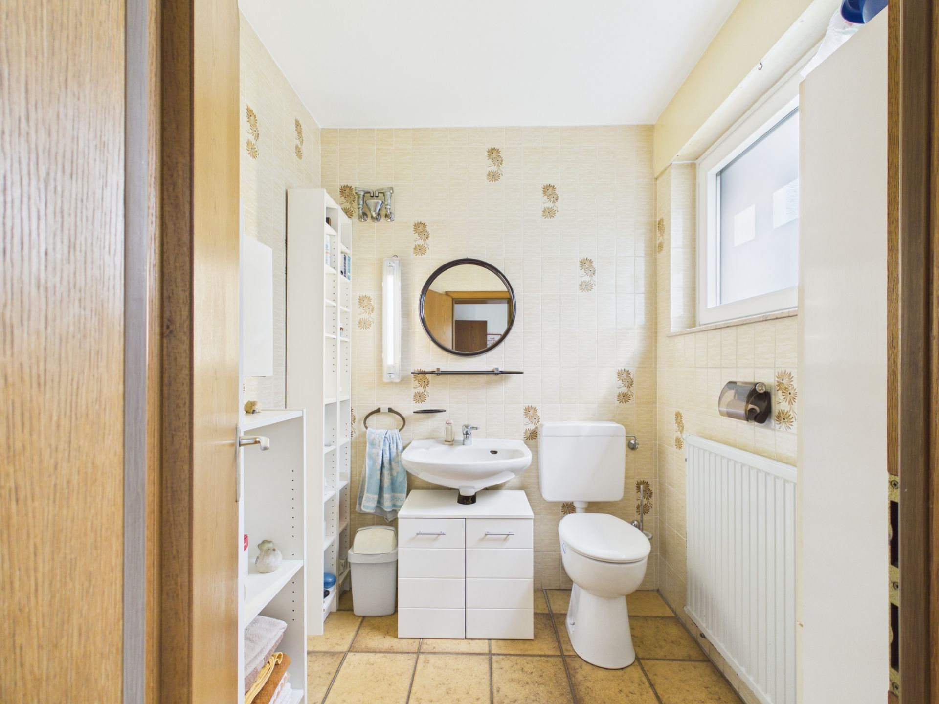 Helles Badezimmer mit Waschbecken, Spiegel, Toilette, Regal und Fenster.