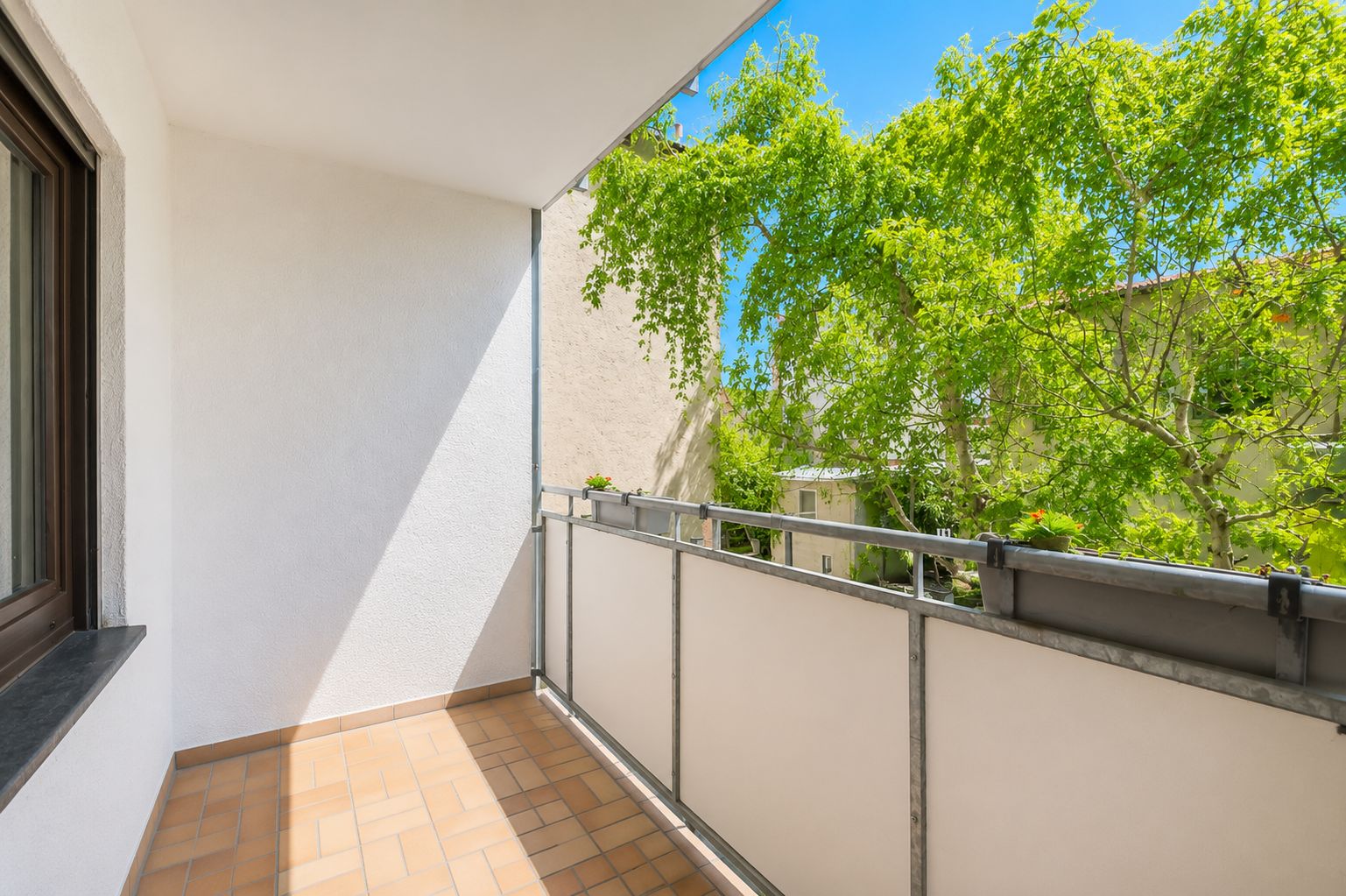 Heller Balkon mit Fliesenboden, Geländer und Blick auf grüne Bäume.