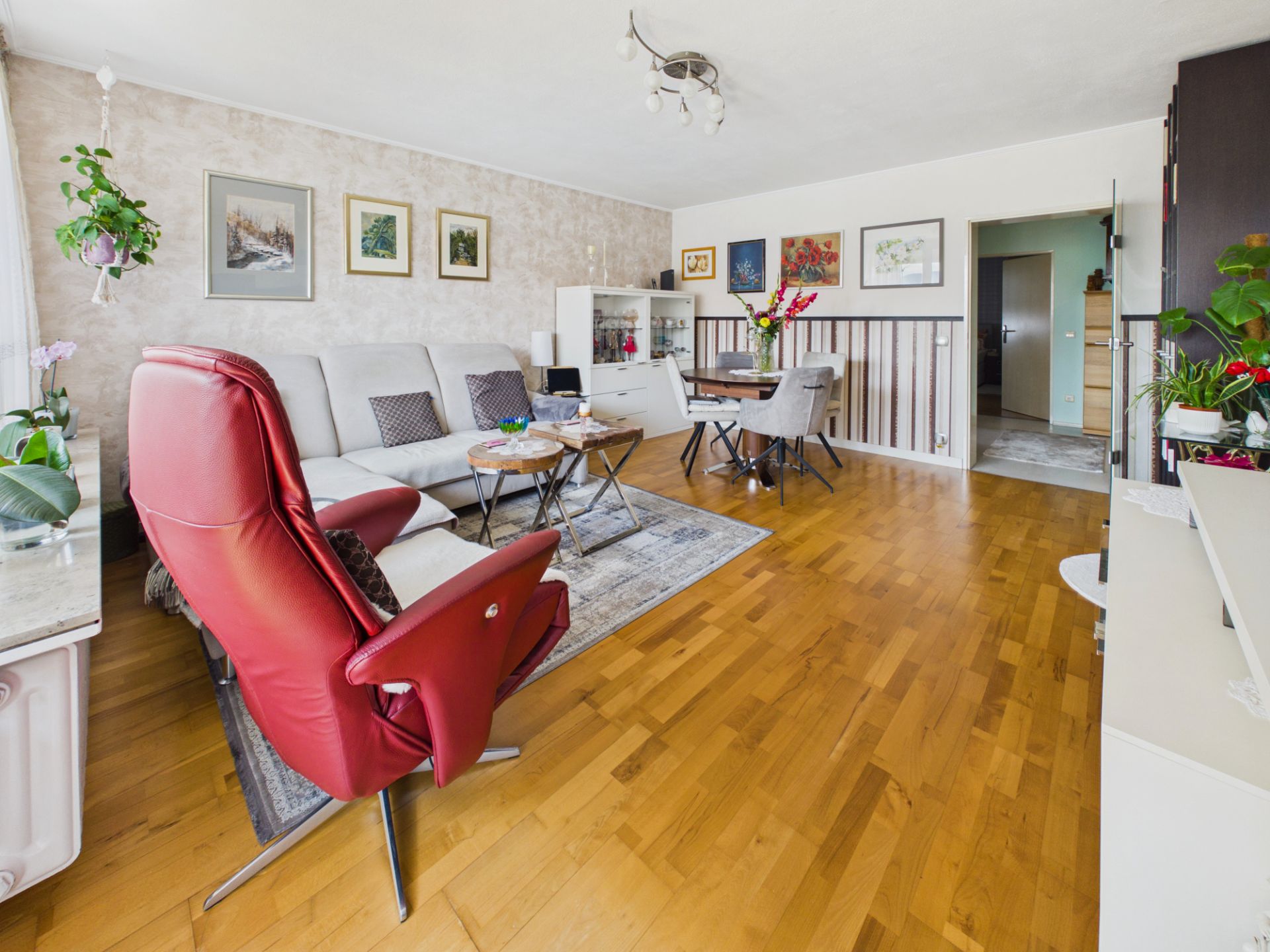 Helles Wohnzimmer mit Holzfußboden, rotem Sessel, grauem Sofa und Esstisch, viele Pflanzen und Bilder an der Wand.