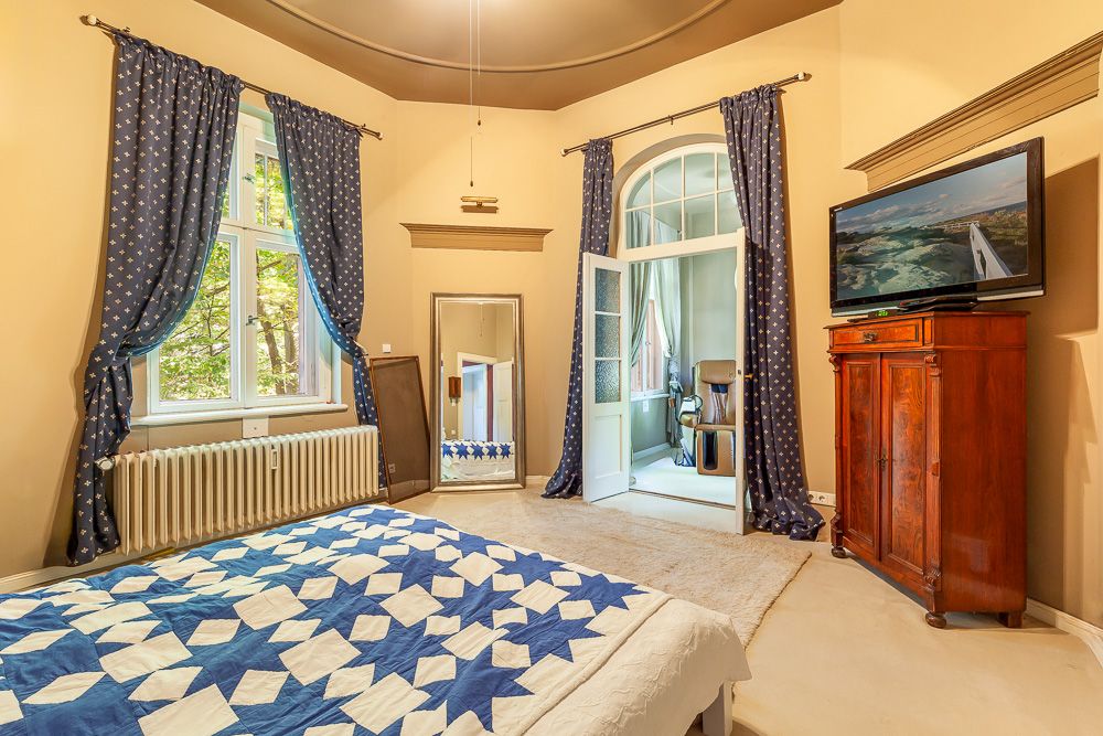 Schlafzimmer mit blau-weißem Bett, Vorhängen, Spiegel, Fernseher und Holzmöbel.
