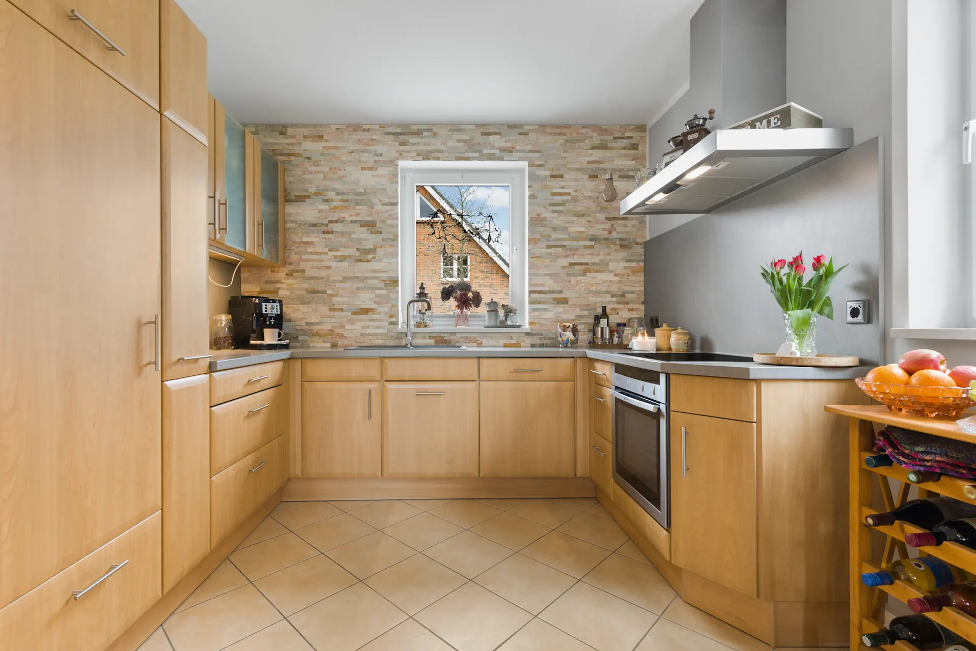 Moderne Küche mit Holzmöbeln, Steintapete, Fenster, Blumen, Obstschale und Weinregal.