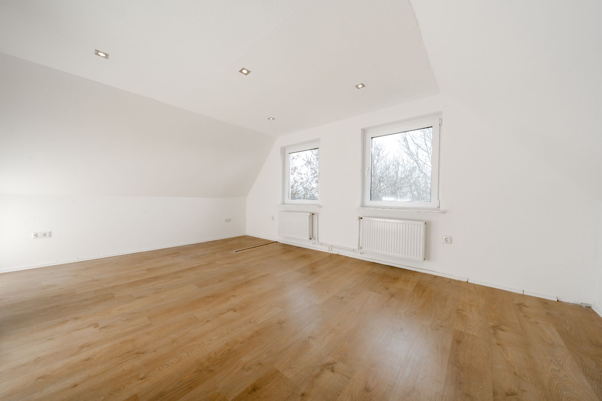 Leerer Raum mit Holzfußboden, weißen Wänden und zwei Fenstern mit Heizkörpern darunter.