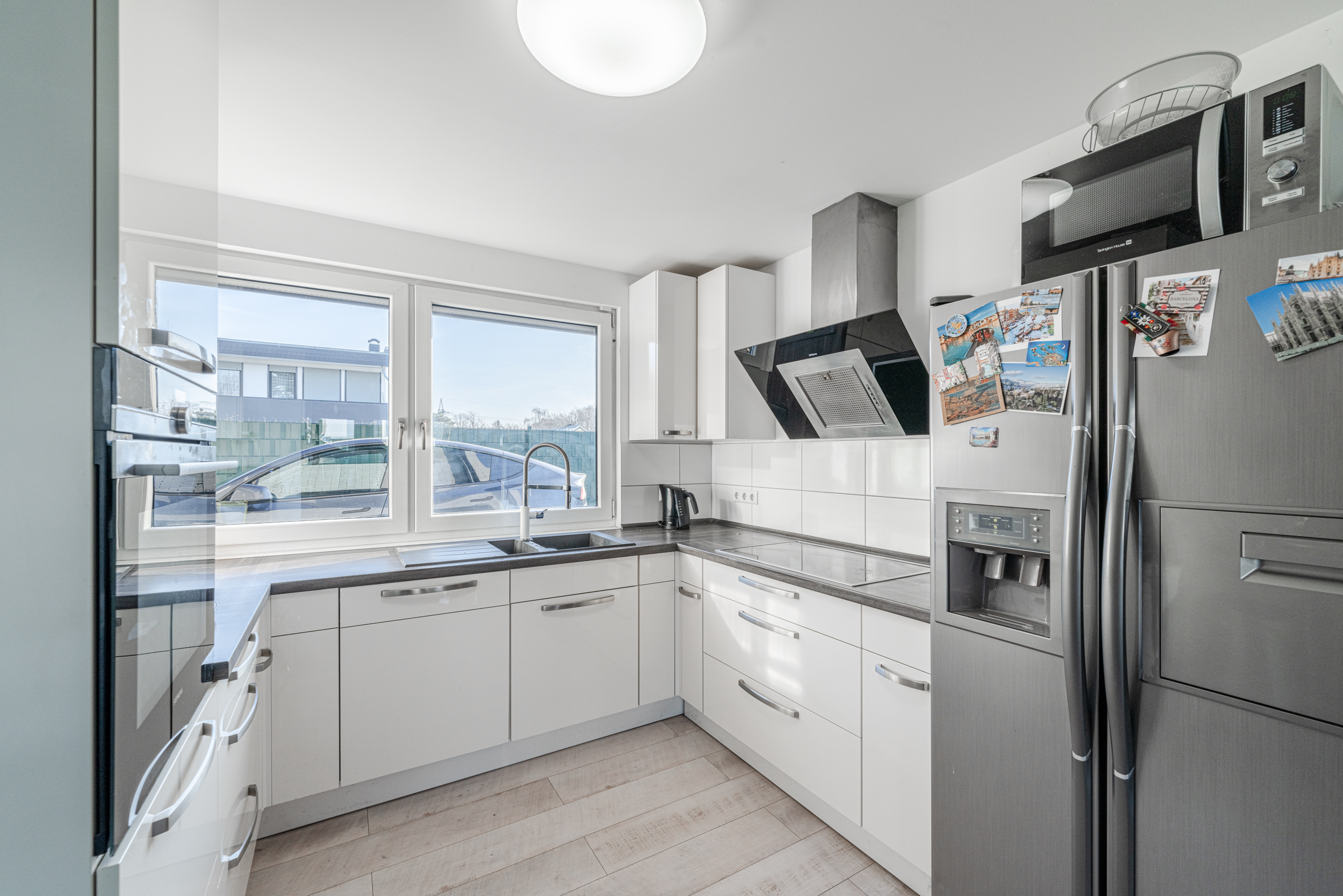 Helle moderne Küche mit weißen Schränken, großem Fenster und Edelstahlkühlschrank.