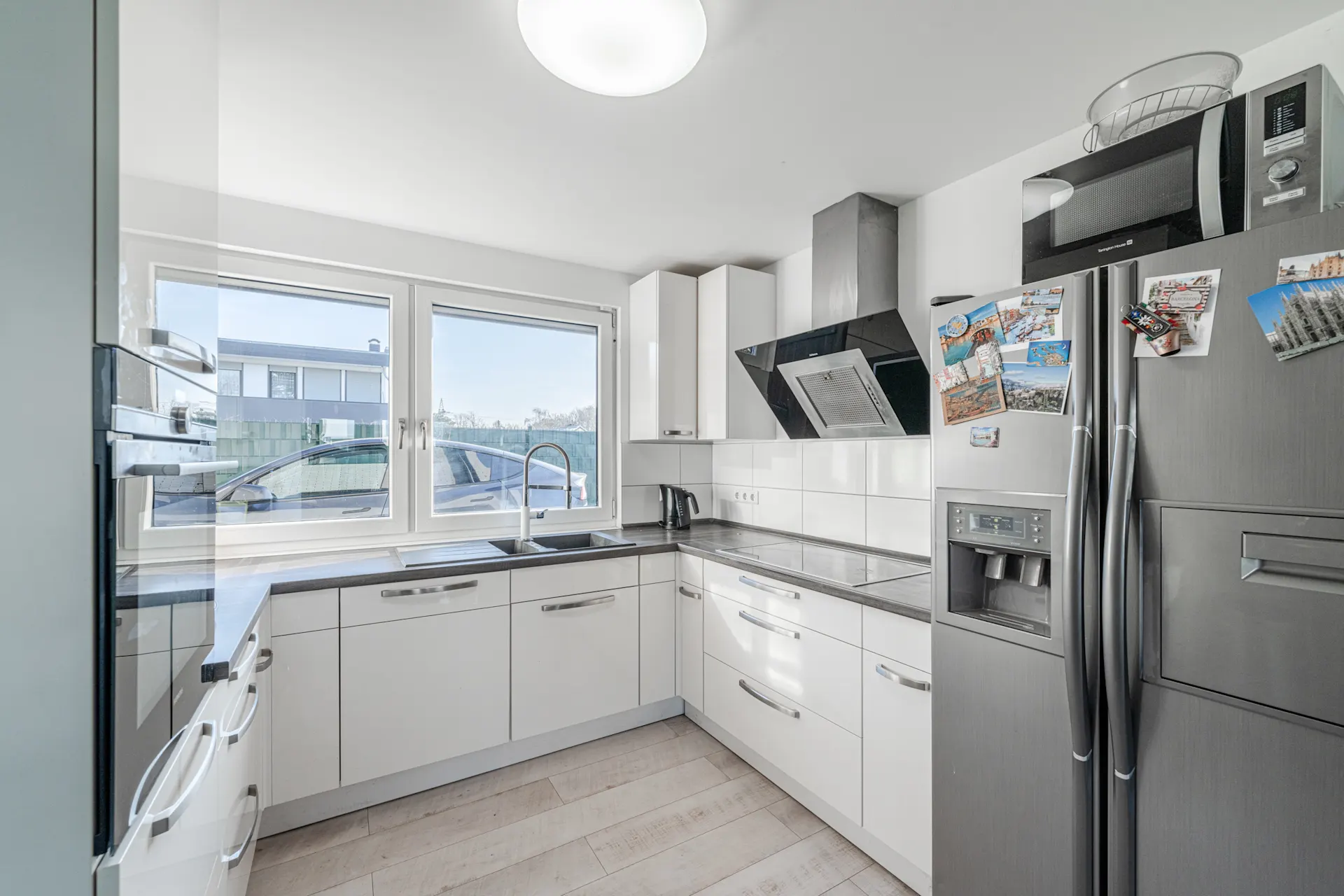 Helle moderne Küche mit weißen Schränken, großem Fenster und Edelstahlkühlschrank.