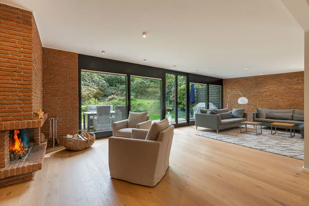 Helles Wohnzimmer mit Kamin, zwei Sofas, Sessel und großen Fenstern zum Garten.