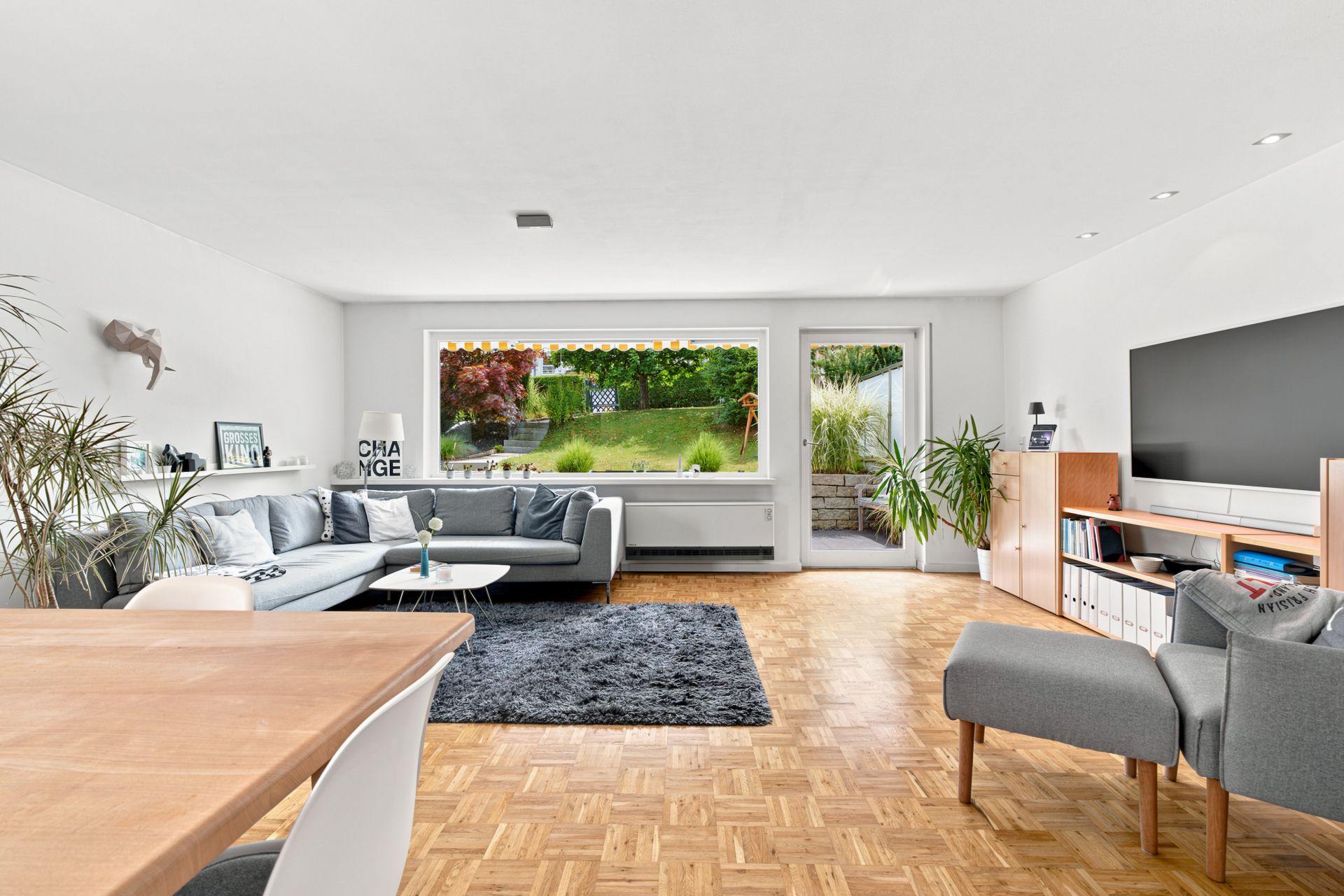 Helles Wohnzimmer mit Parkettboden, Sofa, Sessel, großem Fenster und Zugang zum Garten.