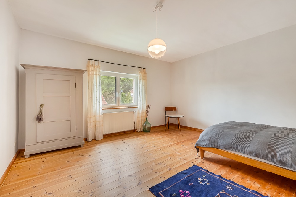 Helles Schlafzimmer mit Holzboden, Bett, Schrank, Tisch und Stuhl am Fenster.