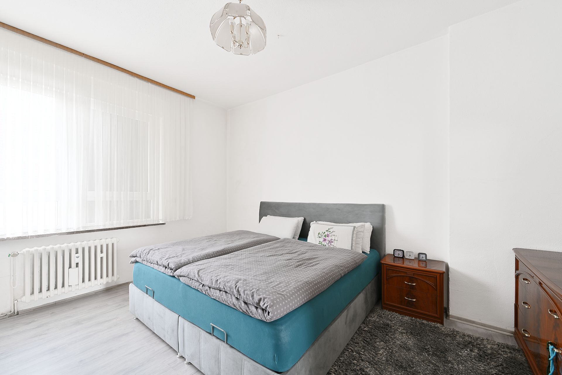 Schlafzimmer mit Doppelbett, Nachttisch, Kommode und großem Fenster mit weißen Vorhängen.
