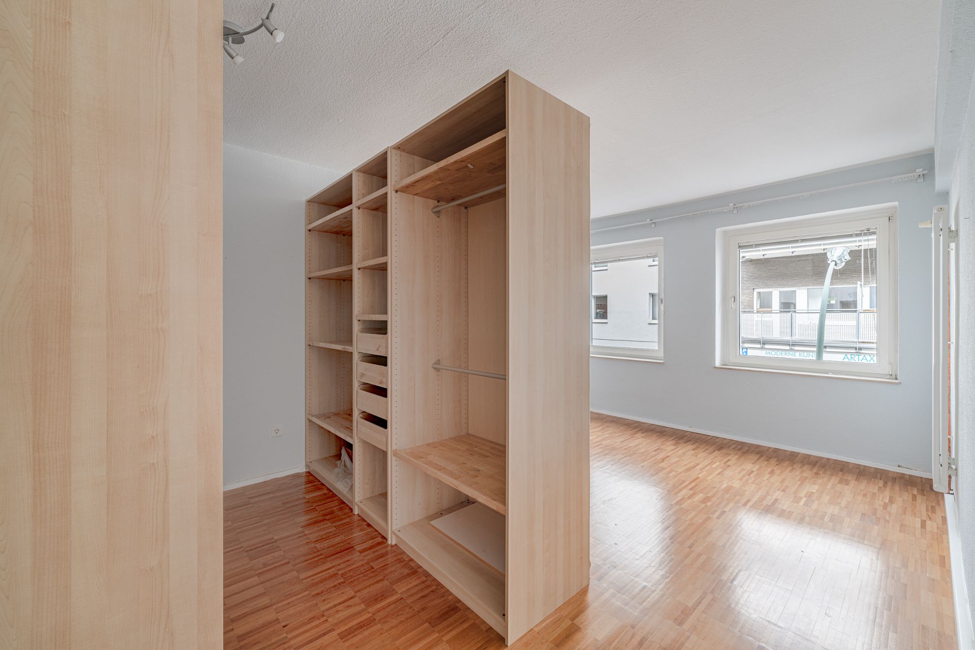 Helles Zimmer mit großen Fenstern und offenem Holzregal auf Parkettboden.