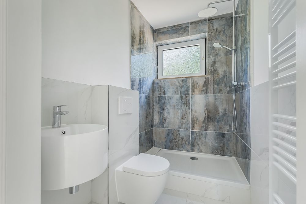 Modernes Bad mit Dusche, WC und kleinem Waschbecken, Fliesenwände und Fenster.