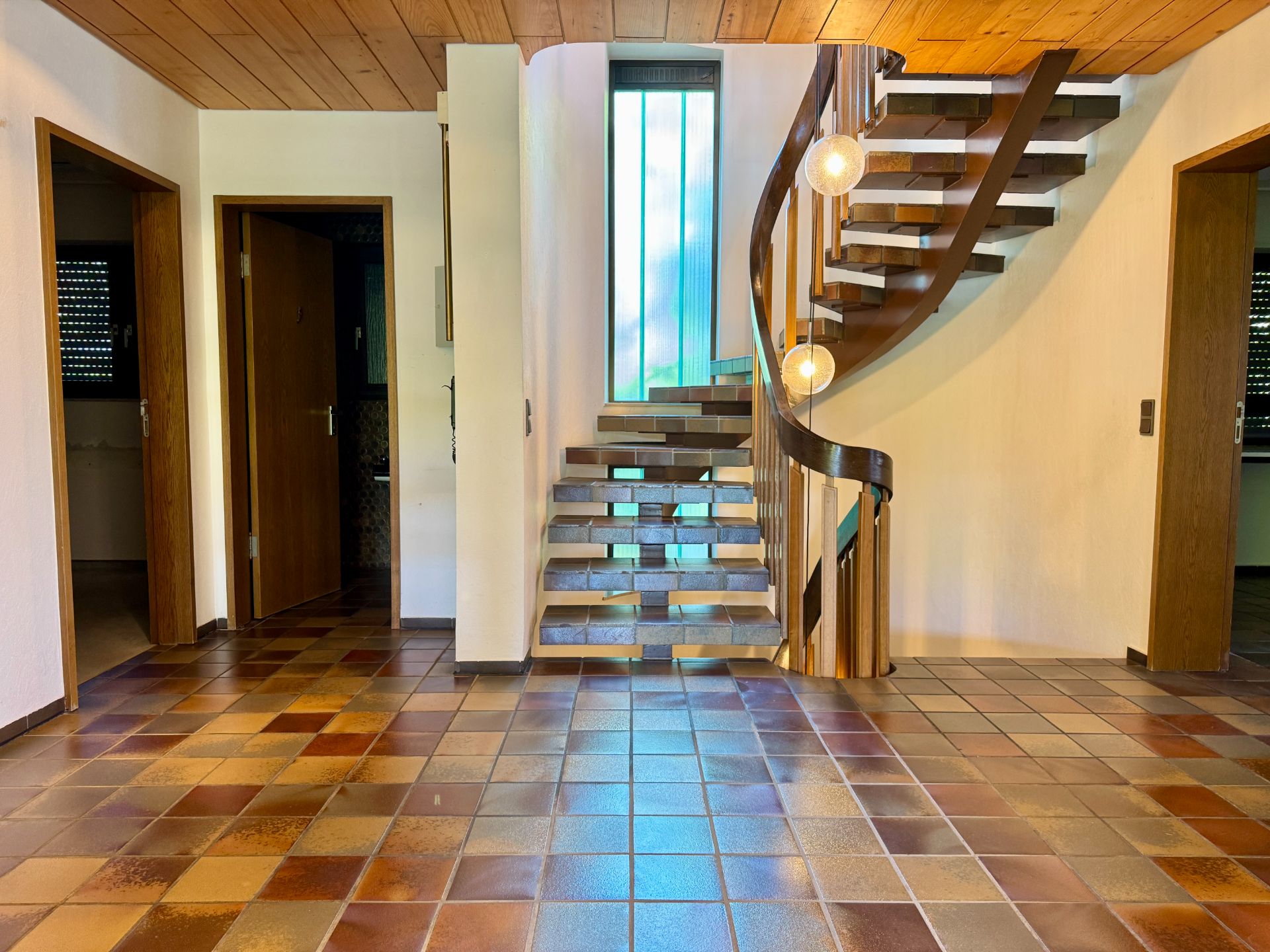 Helles Treppenhaus mit geschwungener Holztreppe und gefliestem Boden in einem Wohnhaus.