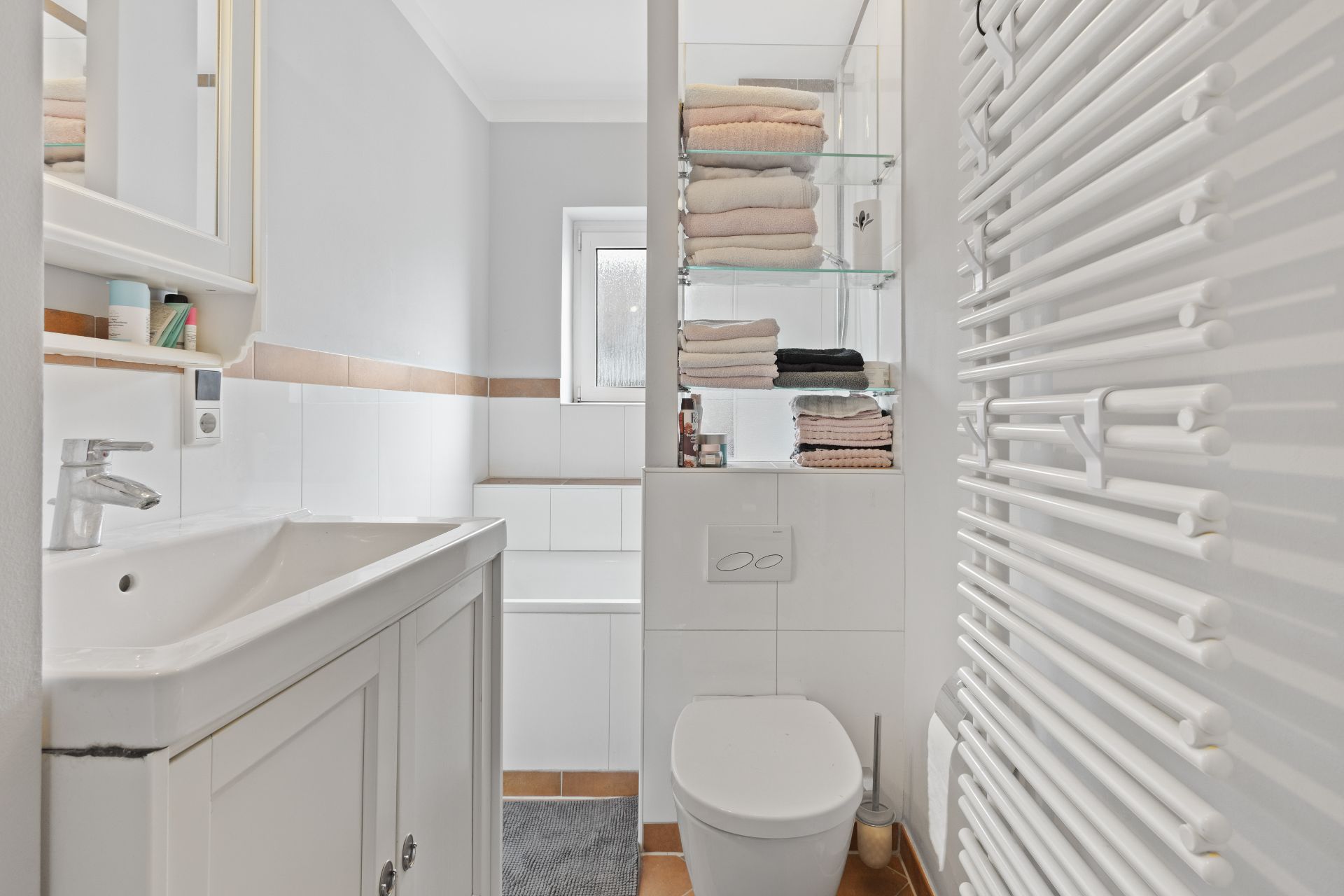 Helles Badezimmer mit Waschbecken, WC, Handtuchheizkörper und Regal mit gefalteten Handtüchern.
