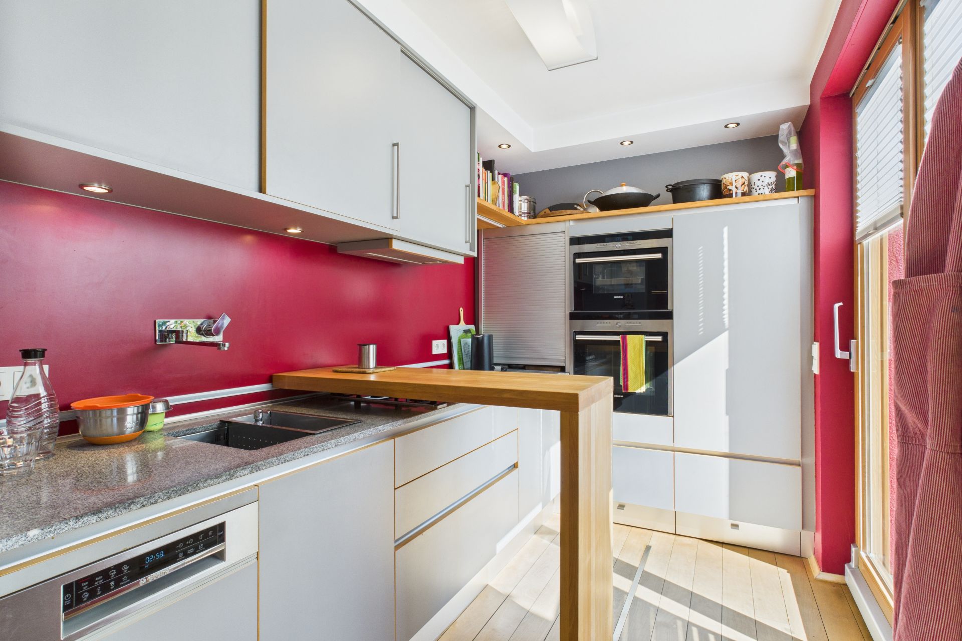 Moderne Küche mit roten Wänden, hellen Schränken, Holztisch und großen Fenstern.