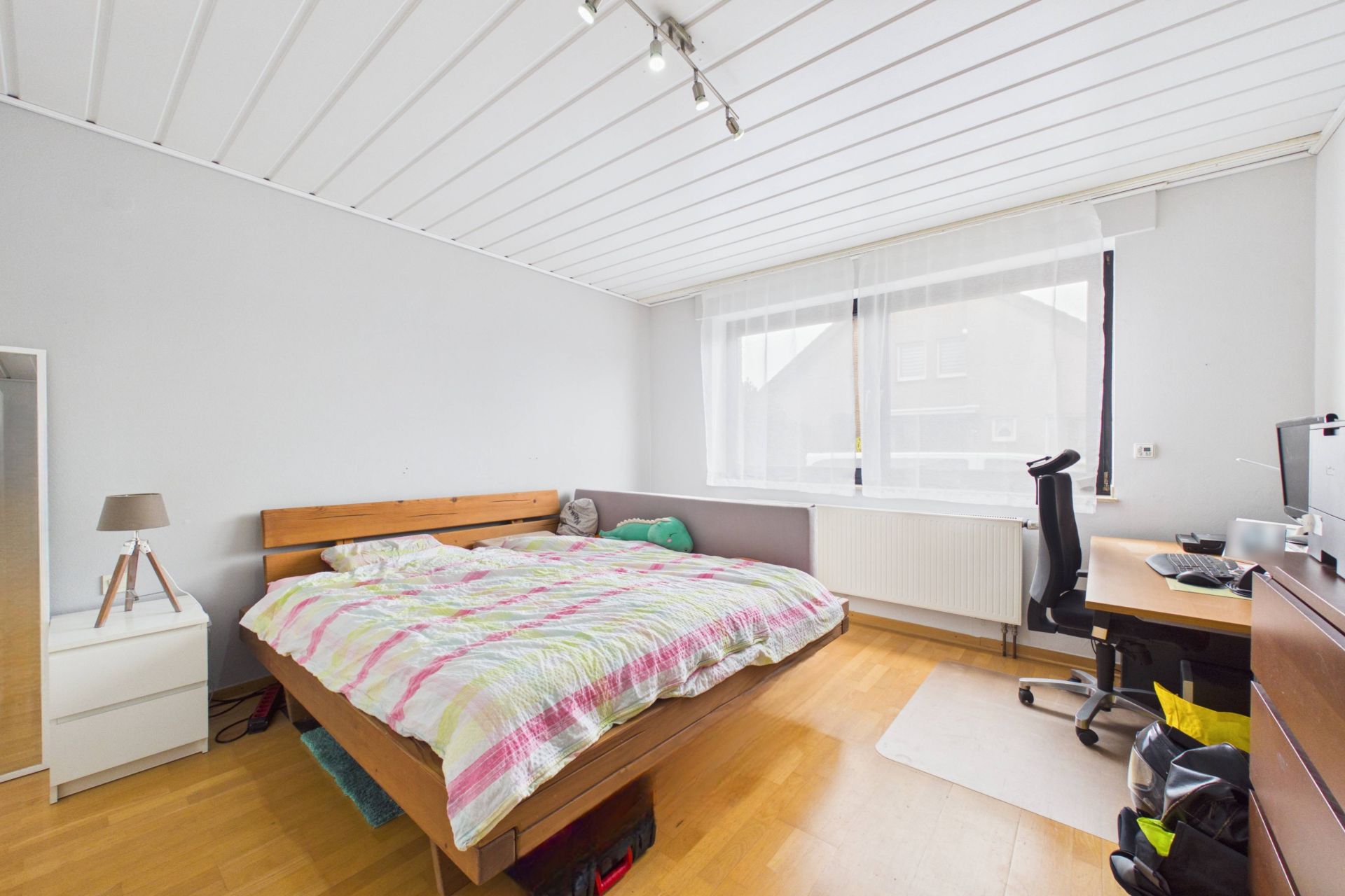 Helles Schlafzimmer mit großem Bett, Schreibtisch, Stuhl und Fenster mit Vorhängen.