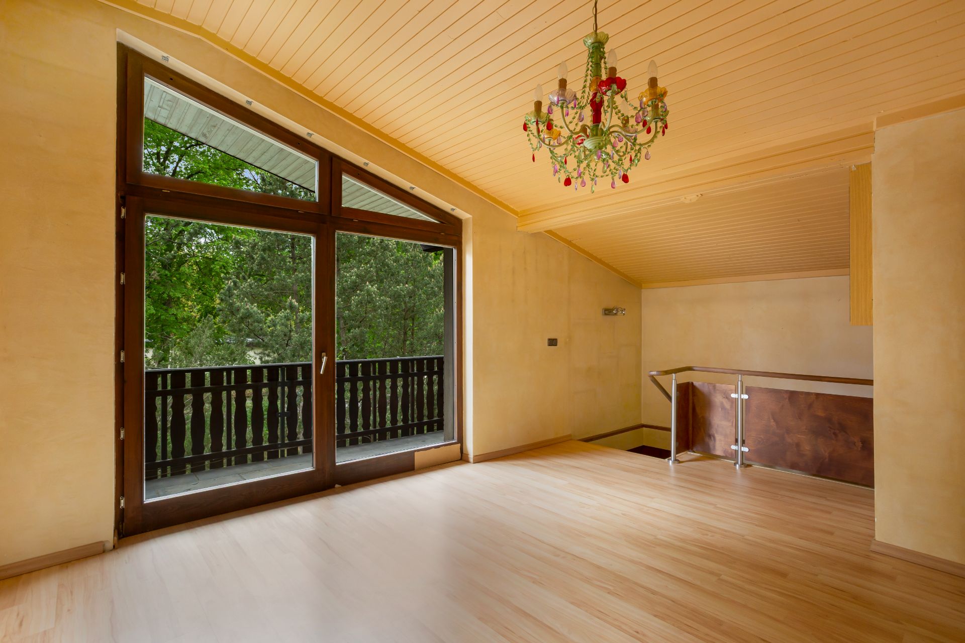 Leerer Raum mit Holzdecke, großem Fenster und Tür zum Balkon, bunter Kronleuchter an der Decke.
