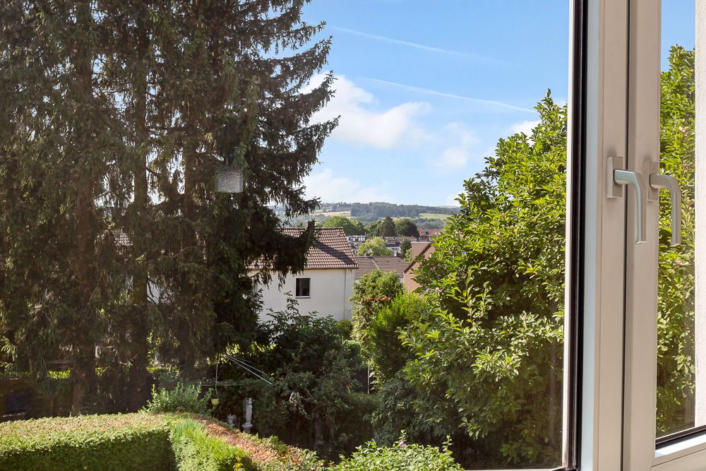 Blick aus einem geöffneten Fenster auf Bäume, Häuser und eine grüne Landschaft.