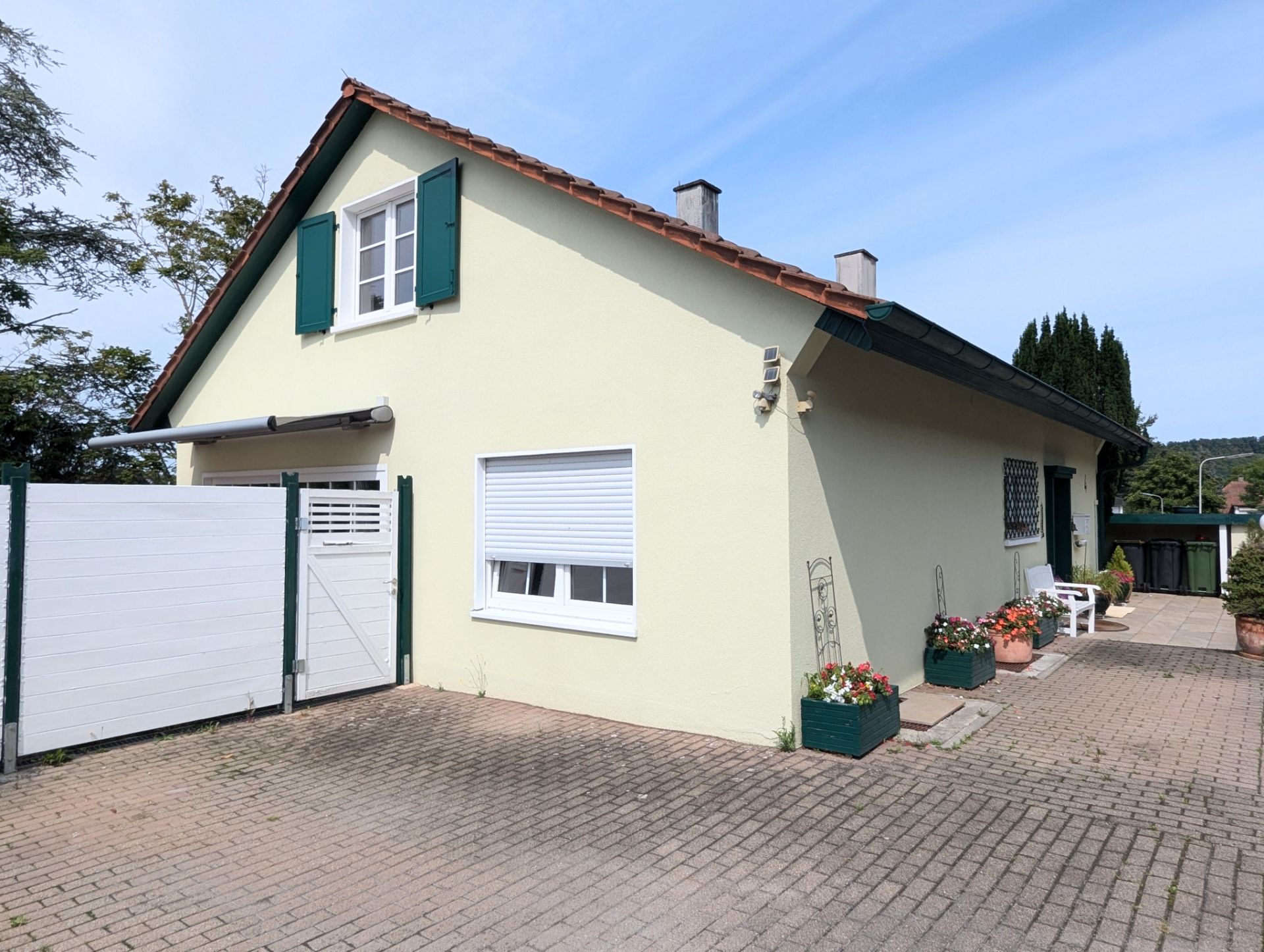 Helles Einfamilienhaus mit grünem Dach, Fensterläden und gepflastertem Hof.