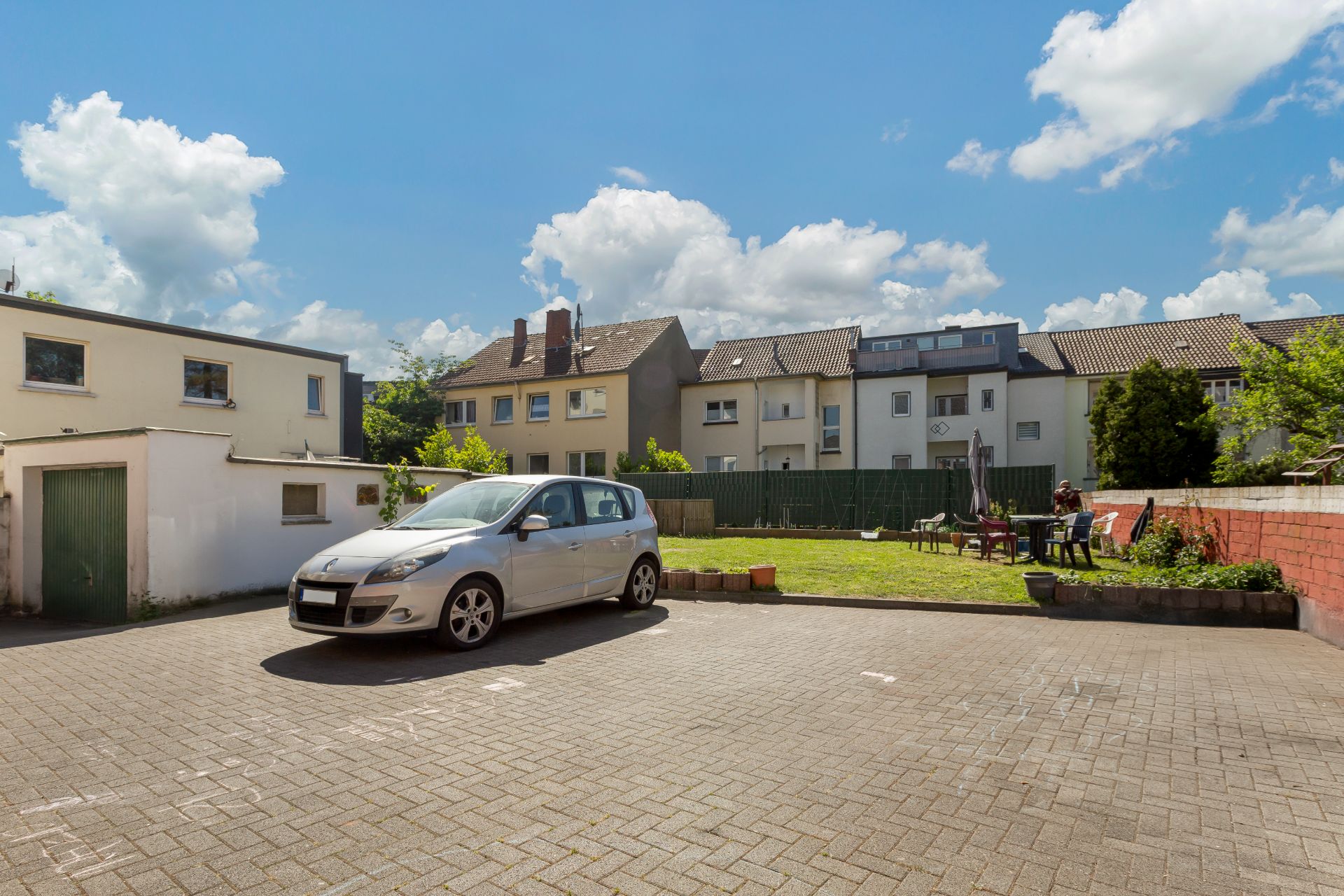 Gepflasterter Parkplatz mit silbernem Auto, angrenzender Garten und Wohnhäuser im Hintergrund.