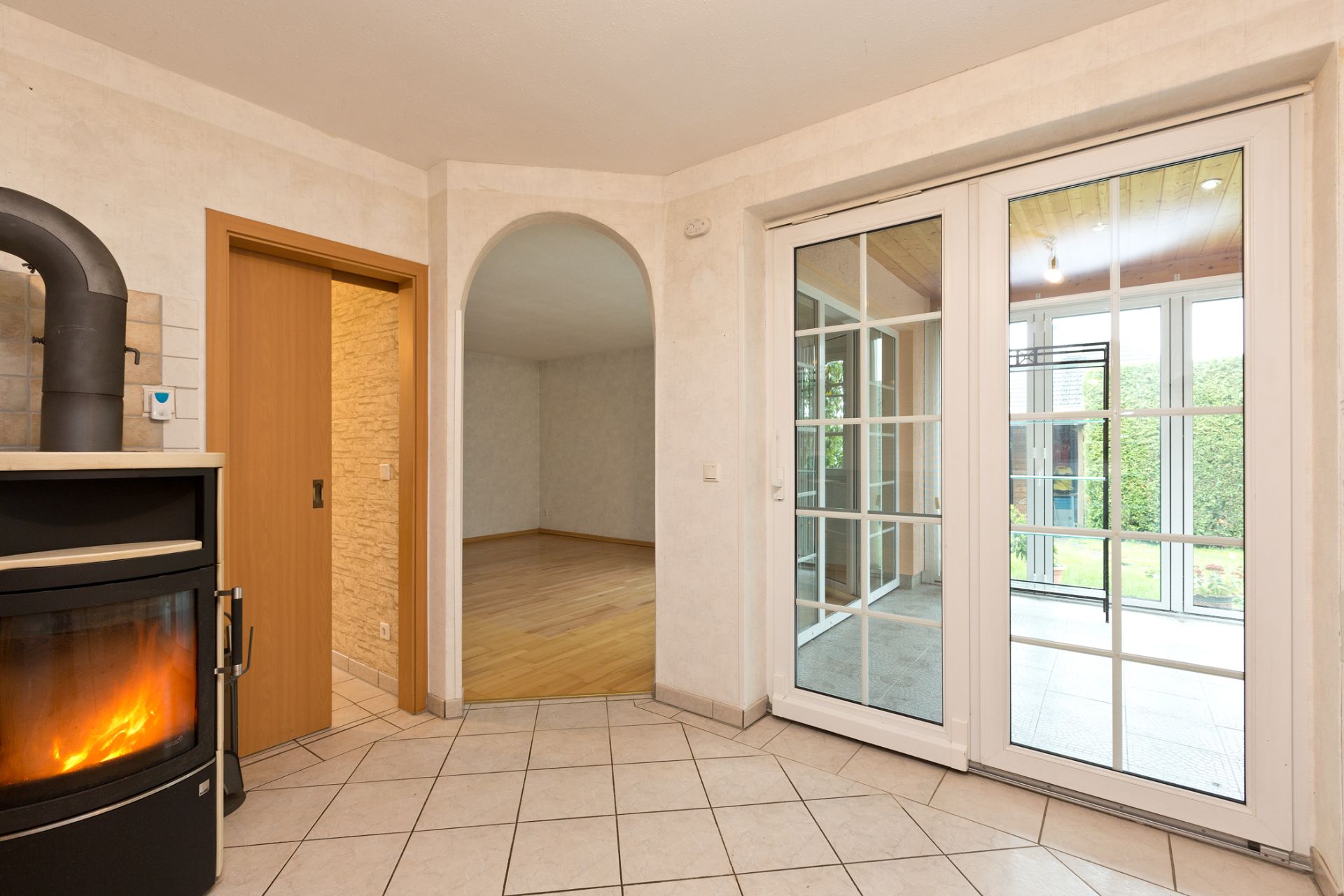 Heller Raum mit Kaminofen, Fliesenboden, Glastüren und Durchgang zu einem Zimmer mit Parkett.