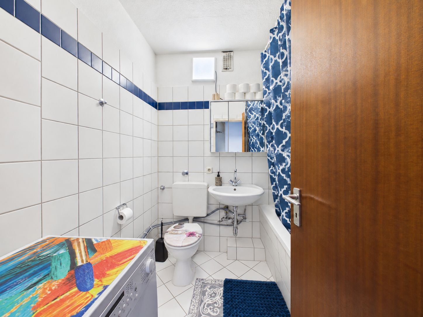 Kleines Badezimmer mit Toilette, Waschbecken, Badewanne und Waschmaschine, weiße Fliesen, blaue Akzente.