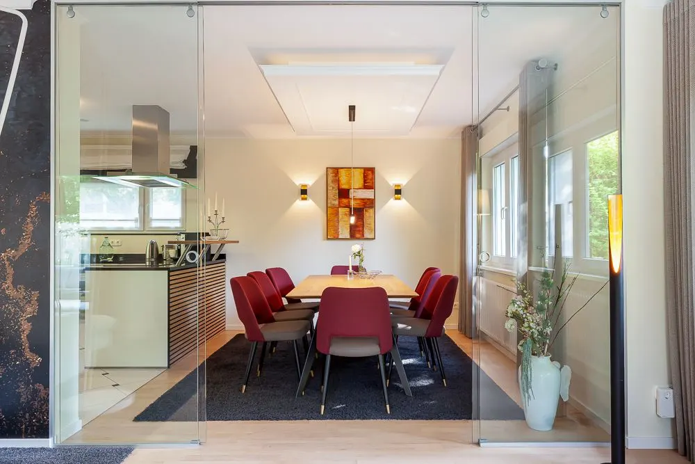 Esszimmer mit Holztisch, acht roten Stühlen und großen Fenstern, daneben moderne Küche.