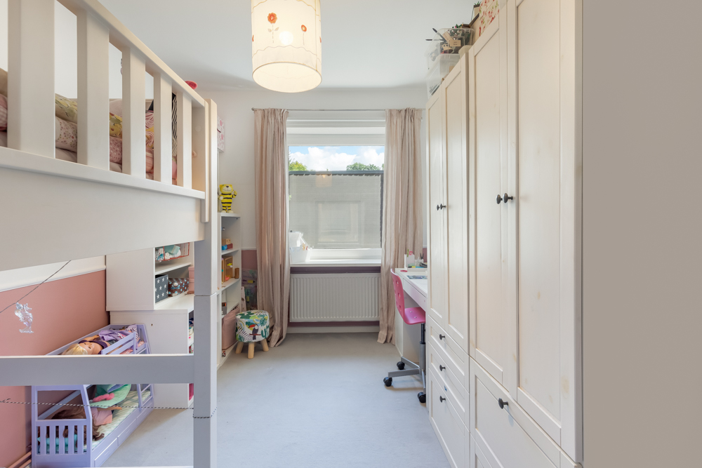 Helles Kinderzimmer mit Hochbett, Schränken, Schreibtisch und Fenster mit Vorhängen.