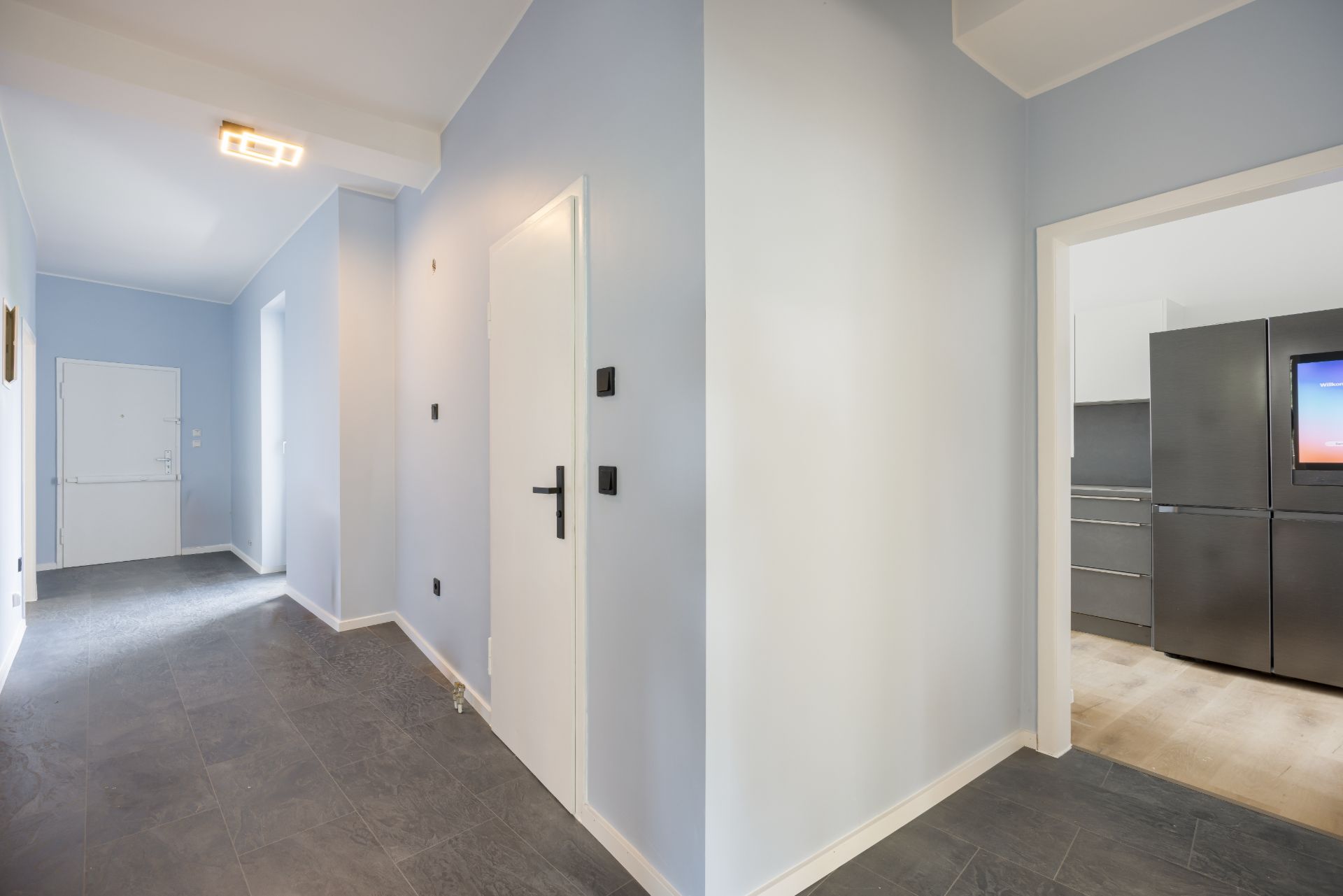 Flur mit hellblauen Wänden, grauem Boden und Blick in eine Küche mit Kühlschrank.