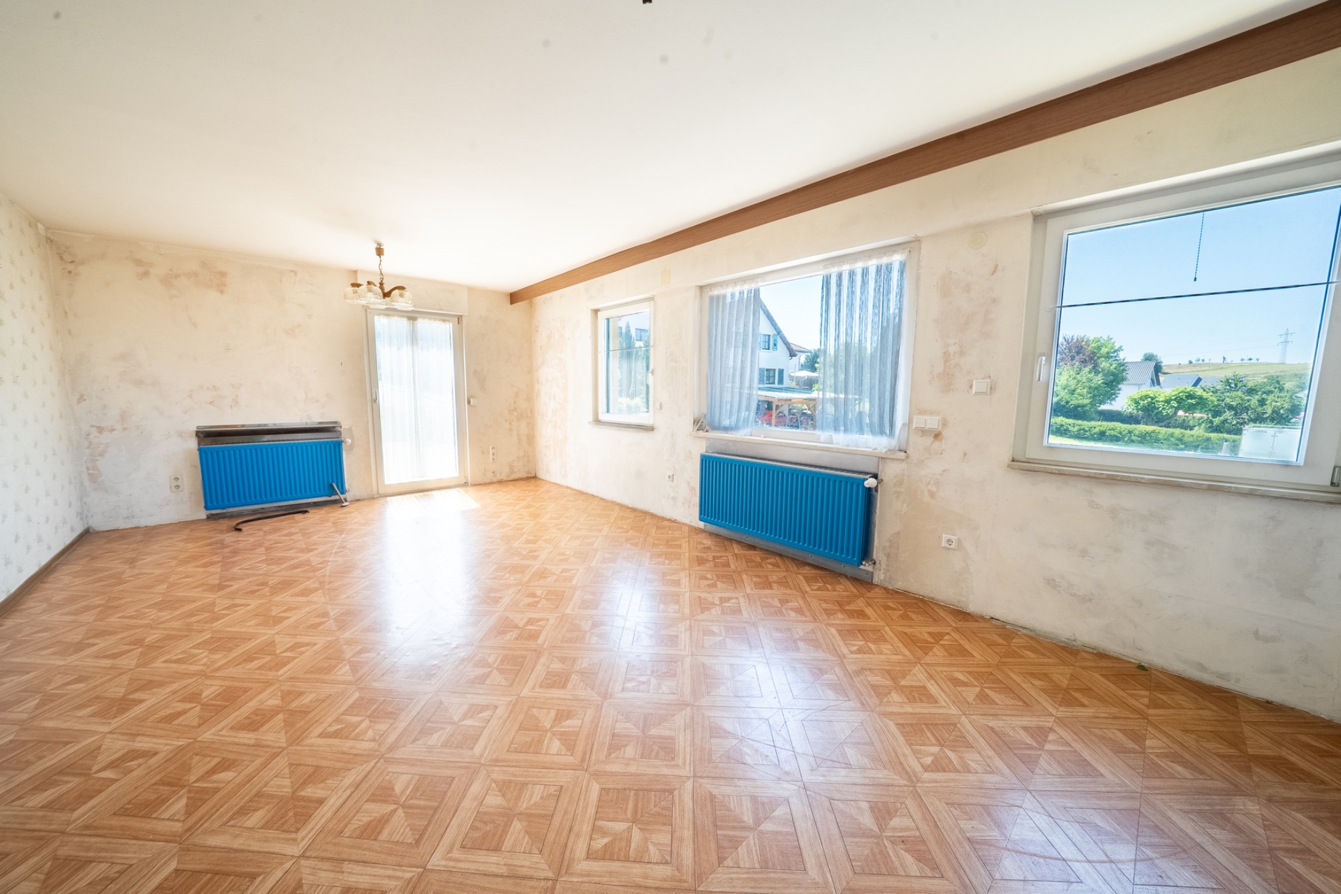 Helles, leeres Zimmer mit Parkettboden, drei Fenstern, zwei blauen Heizkörpern und weißer Wand.