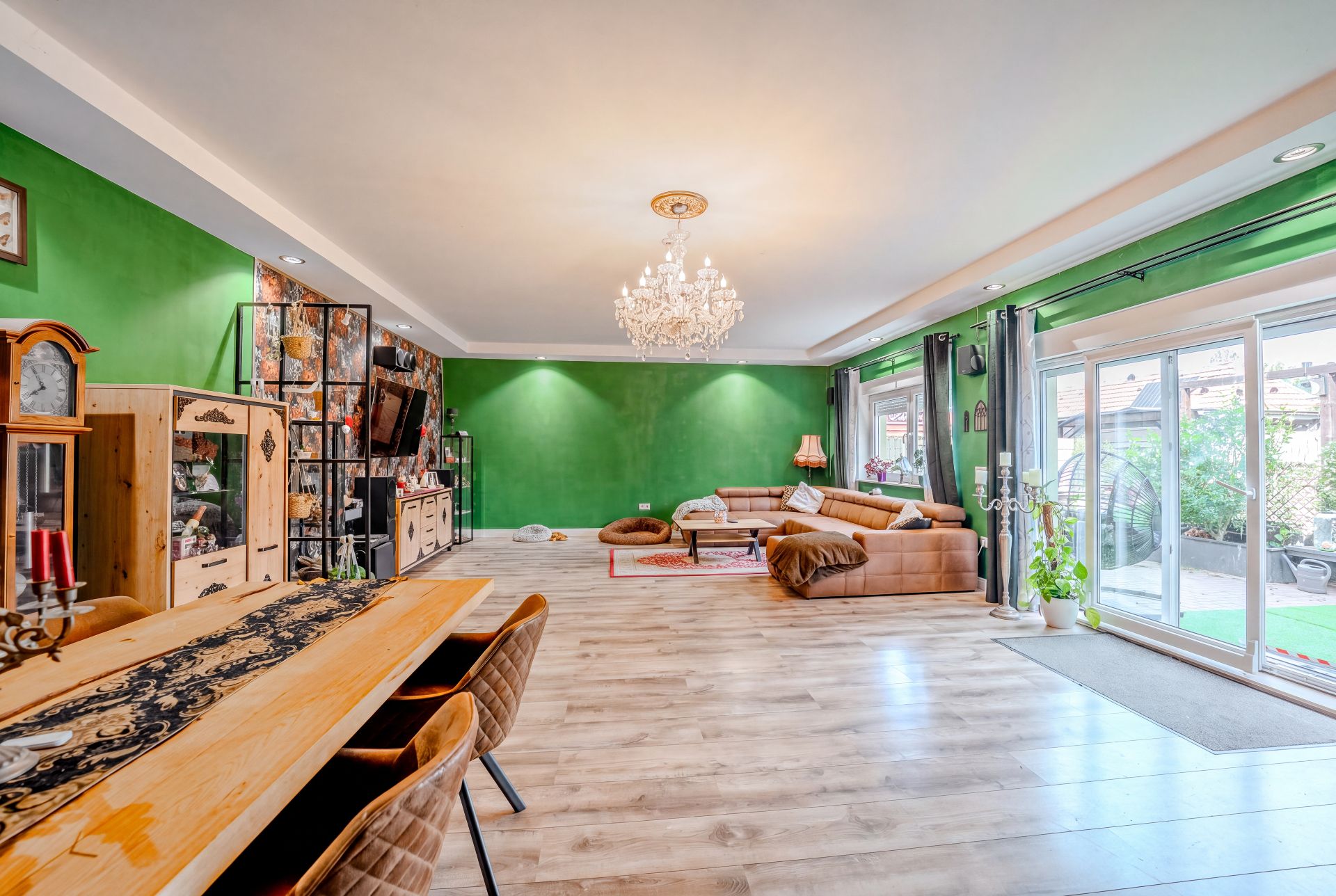 Helles Wohnzimmer mit grünen Wänden, Holzmöbeln, Sofa, Esstisch und großem Fenster zur Terrasse.