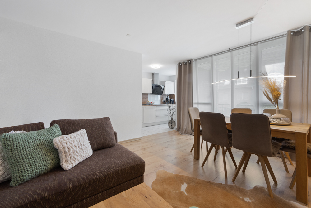 Wohnzimmer mit braunem Sofa, Esstisch, vier Stühlen und offener Küche im Hintergrund.