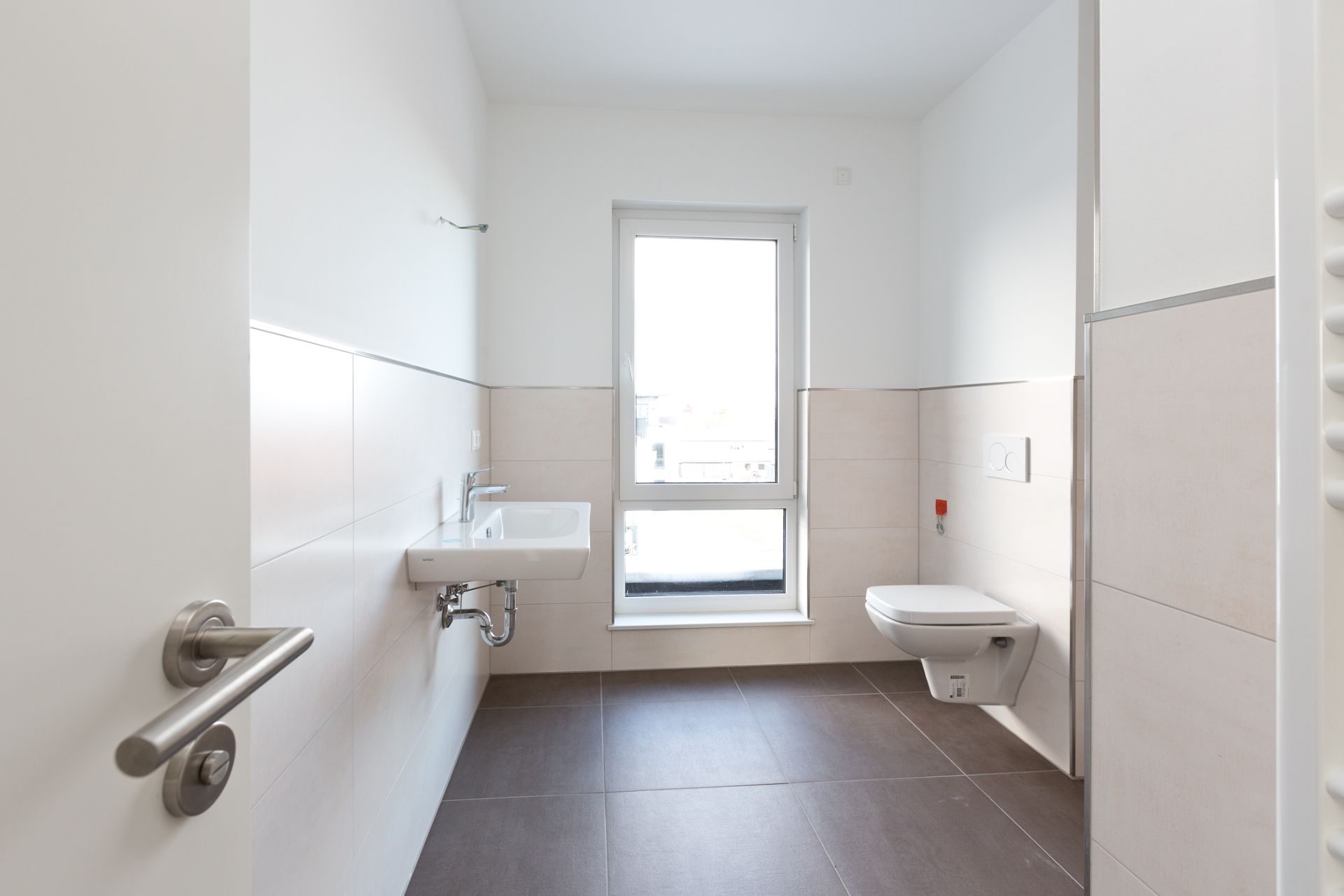 Helles Badezimmer mit Wandwaschbecken, Hänge-WC und Fenster, weiße Wände und große Bodenfliesen.