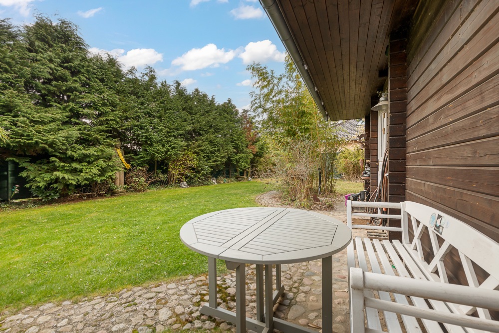 Holzbank und runder Tisch auf Steinterrasse neben Garten mit Rasen und Bäumen.