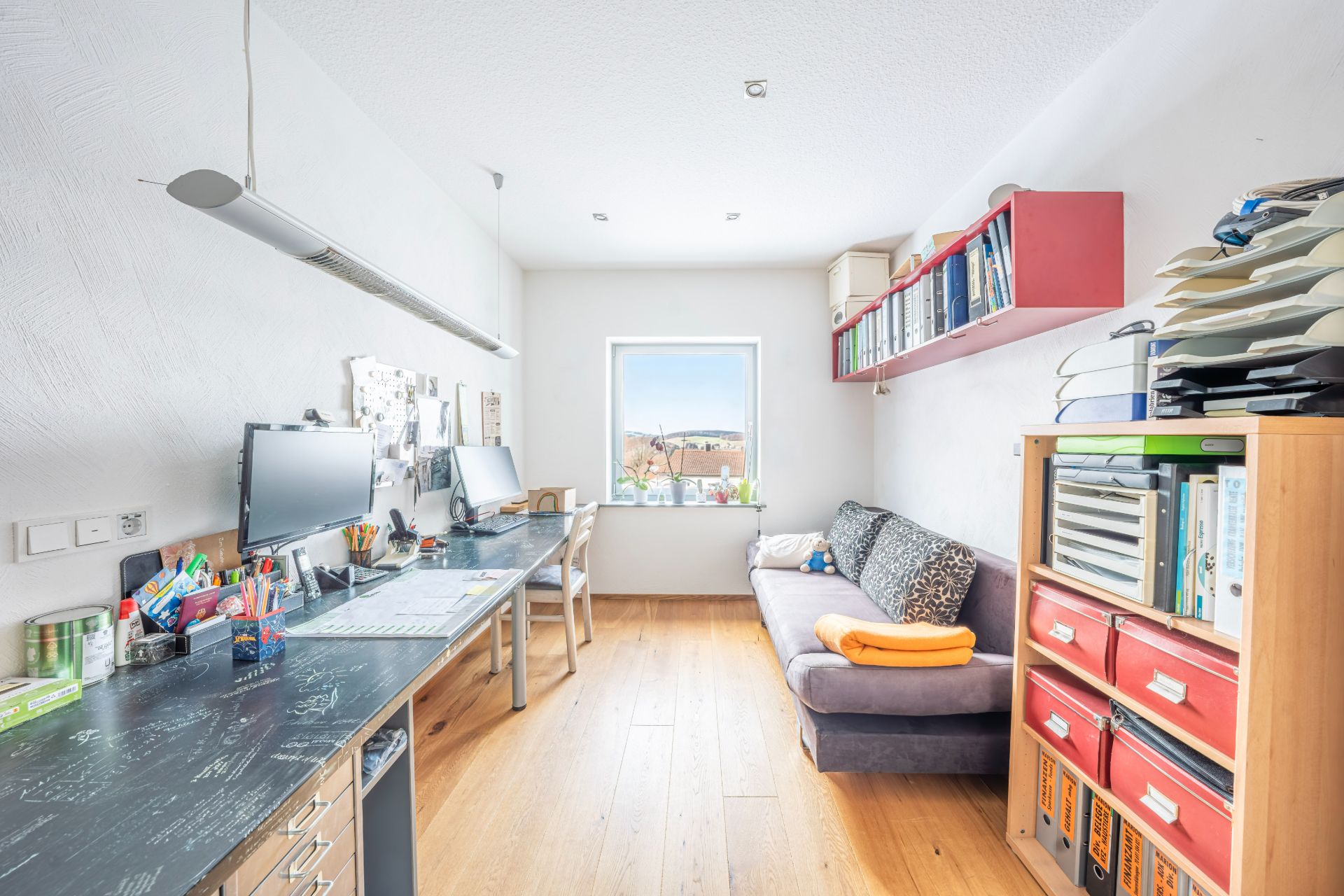 Helles Arbeitszimmer mit Schreibtisch, Computern, Regal und Sofa, Fenster mit Tageslicht.