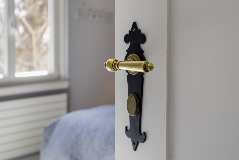 Goldfarbener Türgriff mit schwarzer Platte an einer weißen Tür, im Hintergrund ein Fenster.