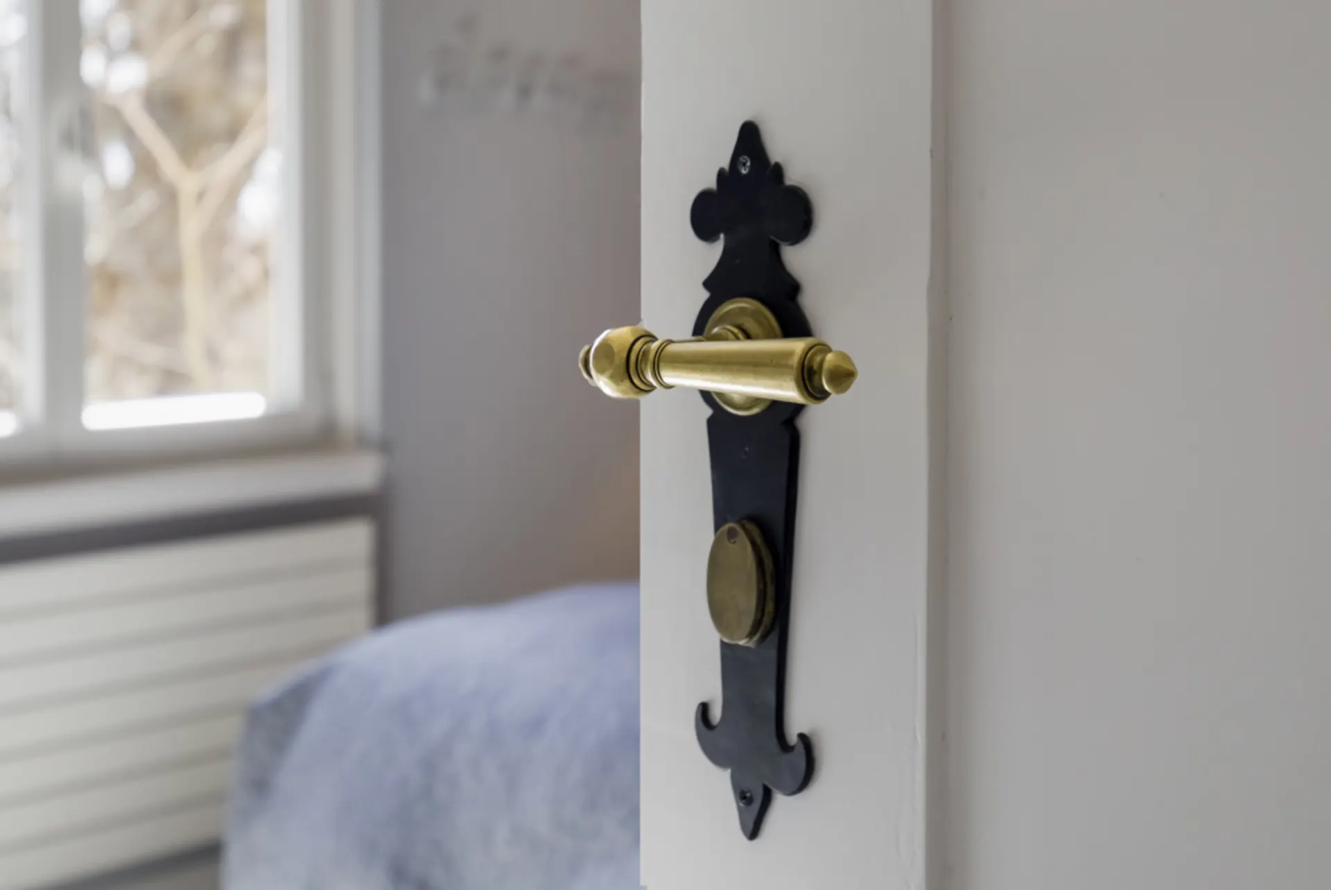 Goldfarbener Türgriff mit schwarzer Platte an einer weißen Tür, im Hintergrund ein Fenster.