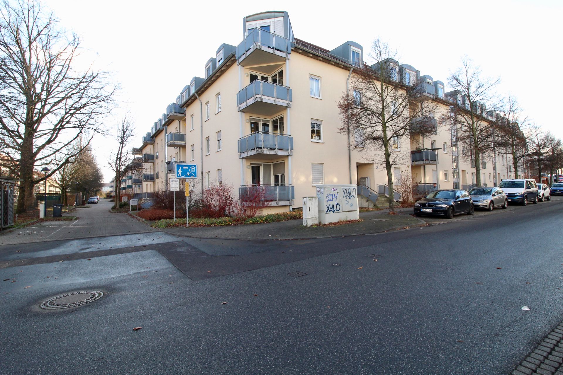 Mehrfamilienhaus an einer Straßenecke mit Balkonen und parkenden Autos.