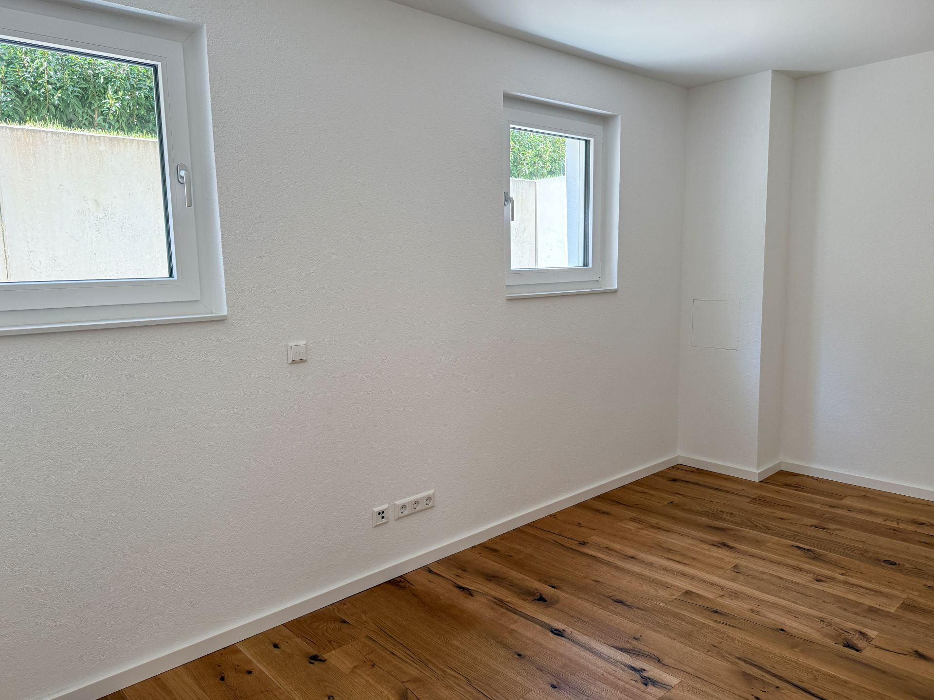 Helles Zimmer mit zwei Fenstern, weißen Wänden und Holzfußboden.