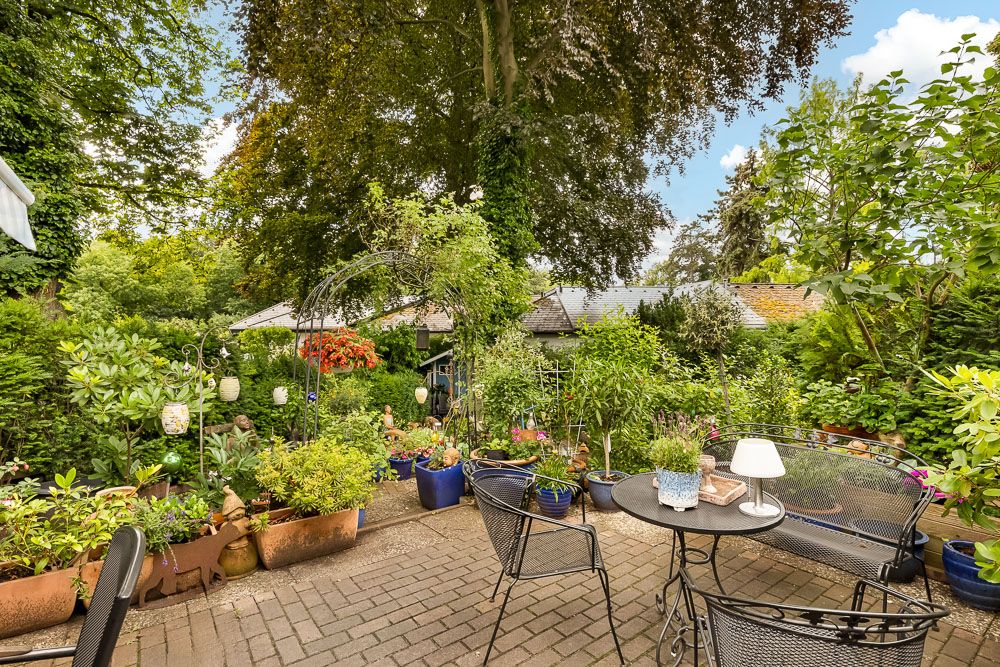 Gepflasterte Terrasse mit Metallmöbeln, vielen Blumentöpfen und üppigem Garten im Hintergrund.