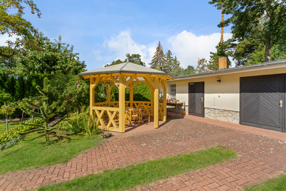 Gelber Pavillon mit Holzmöbeln im Garten, daneben ein Haus mit zwei dunklen Garagentoren.