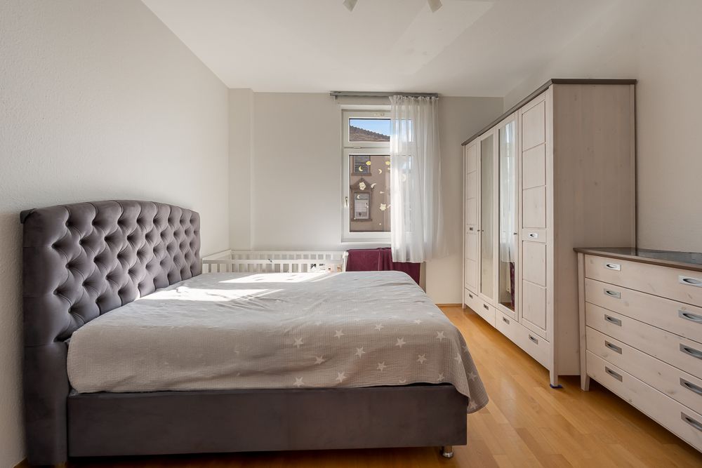 Helles Schlafzimmer mit Doppelbett, Kleiderschrank, Kommode und Fenster mit Vorhang.