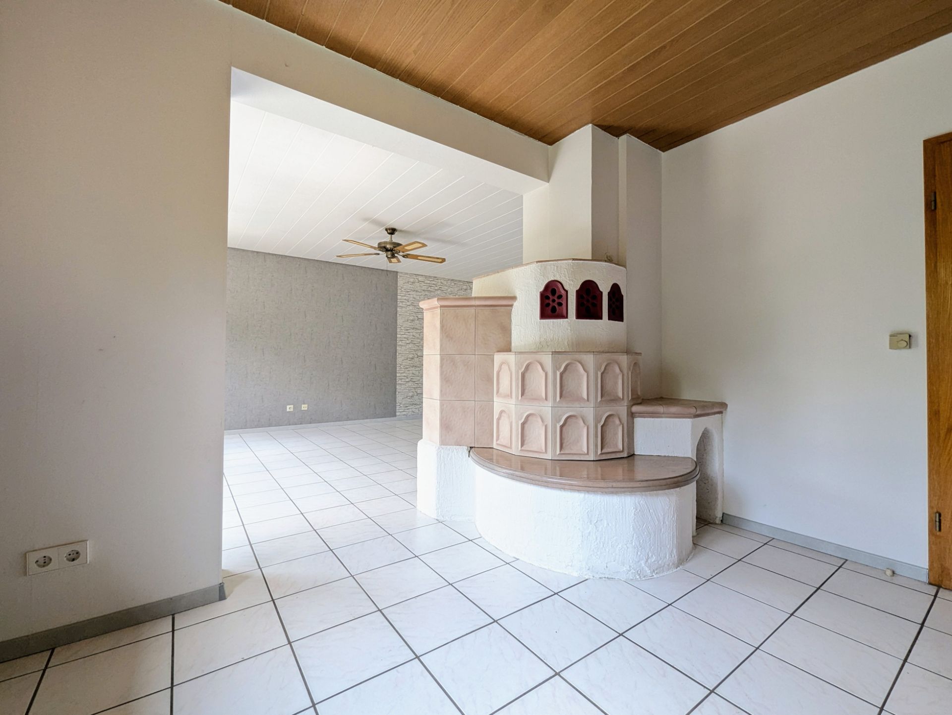 Helles Wohnzimmer mit Kachelofen, weißen Fliesen und Deckenventilator.