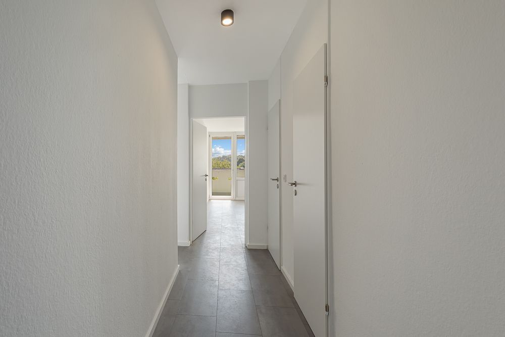 Heller Flur mit grauem Boden, weißen Wänden und mehreren Türen, am Ende ein Fenster mit Ausblick.