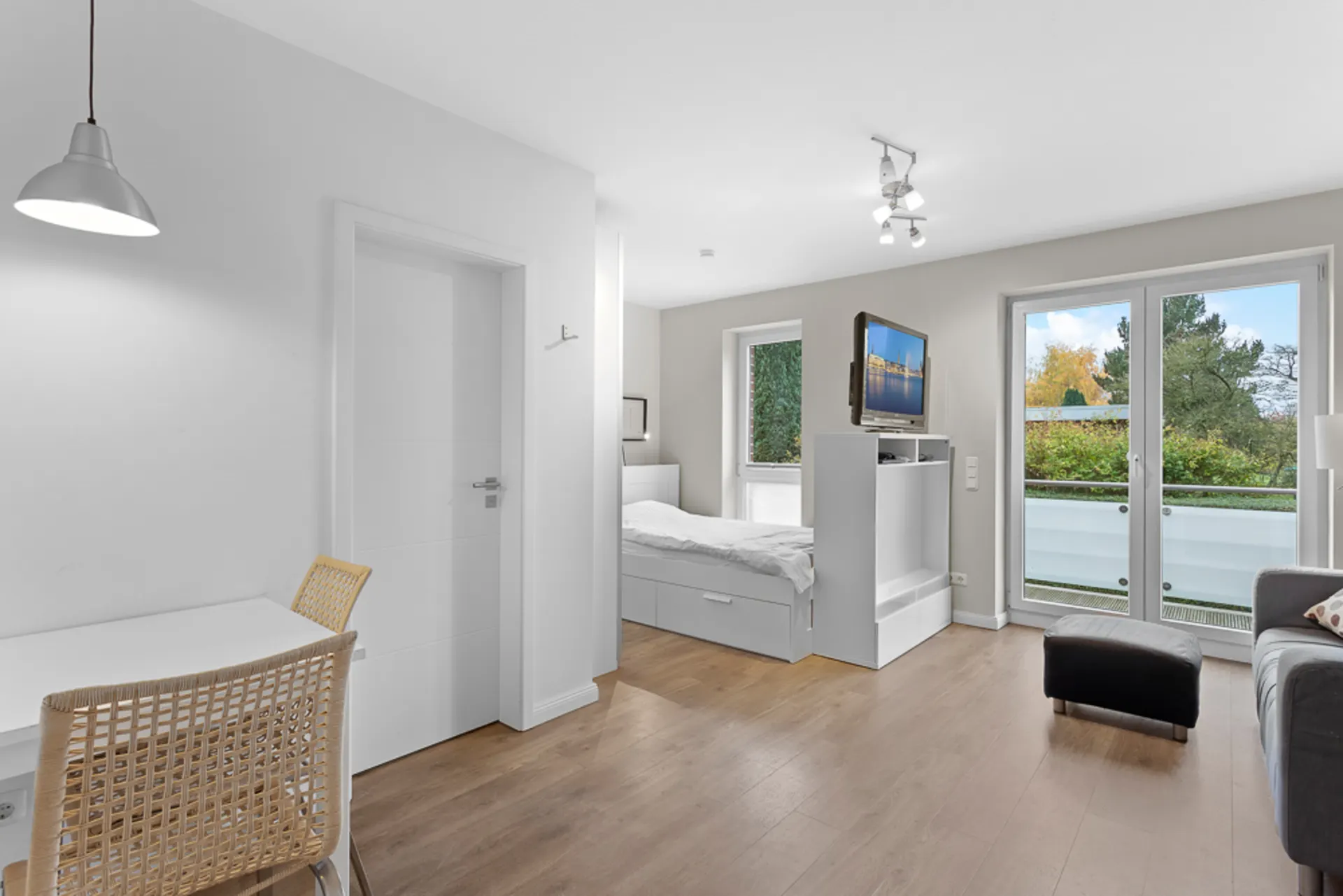 Helles Zimmer mit Bett, Schreibtisch, Fernseher und großen Fenstern mit Blick auf einen Balkon.