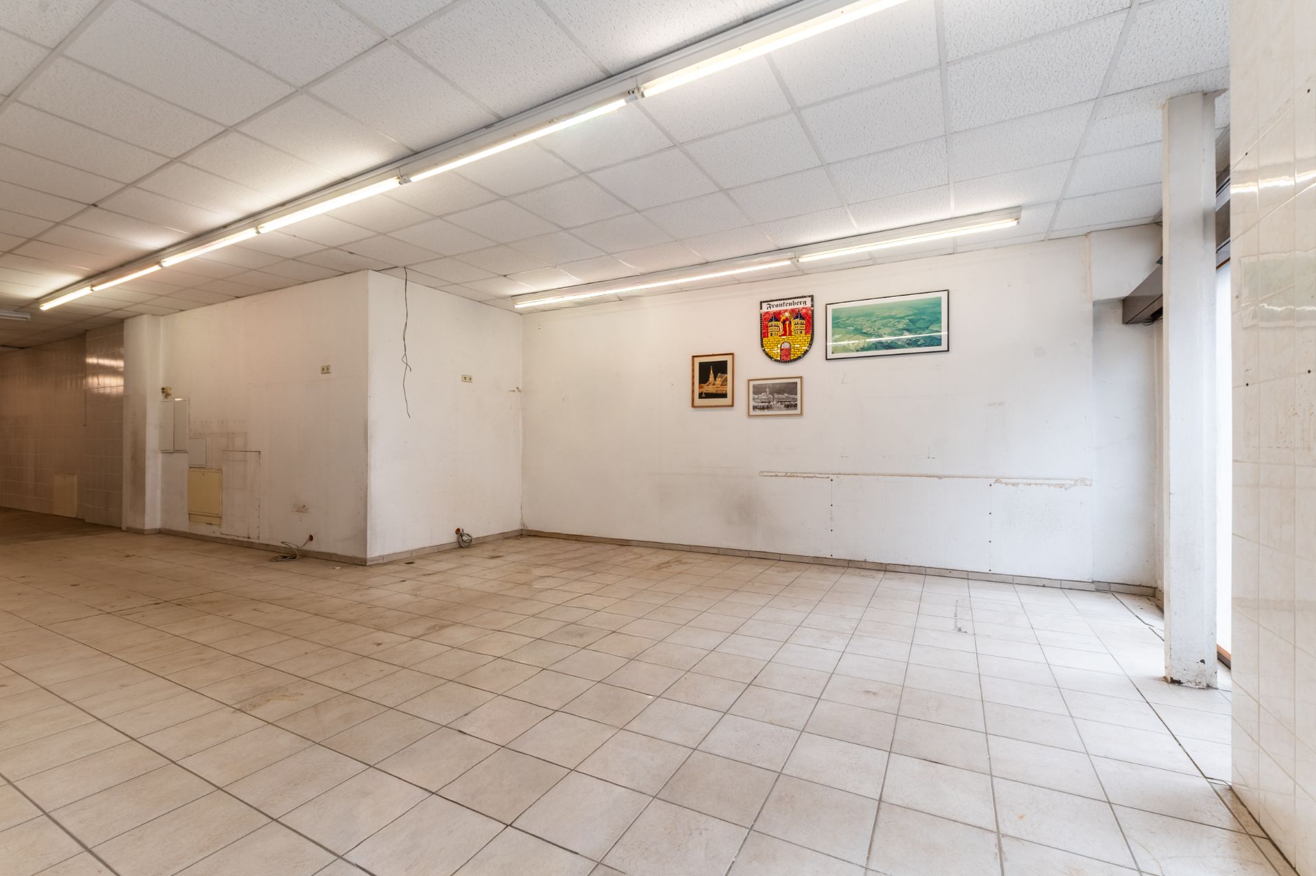 Leerer Raum mit Fliesenboden, weißer Decke und einigen Bildern an der Wand.