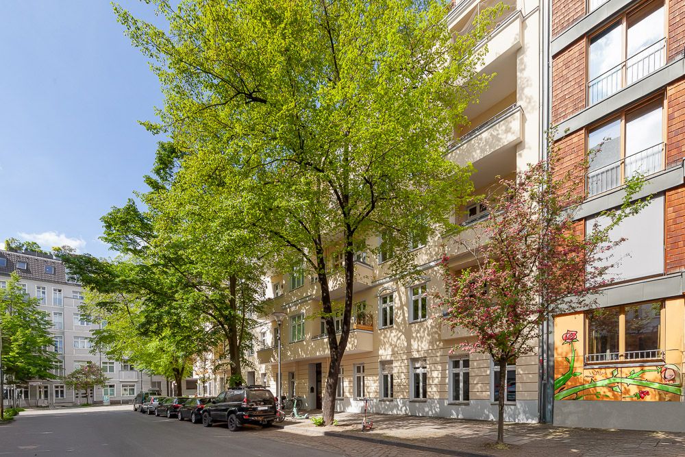 Baumbestandene Wohnstraße mit mehrstöckigen Gebäuden und parkenden Autos bei Tageslicht.