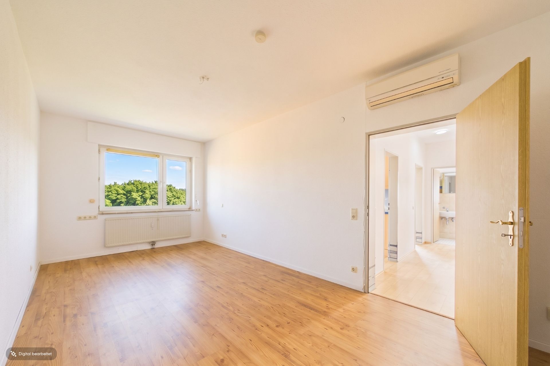Leeres Zimmer mit Holzfußboden, großem Fenster und Klimaanlage, Tür öffnet zu Flur mit Blick ins Bad.